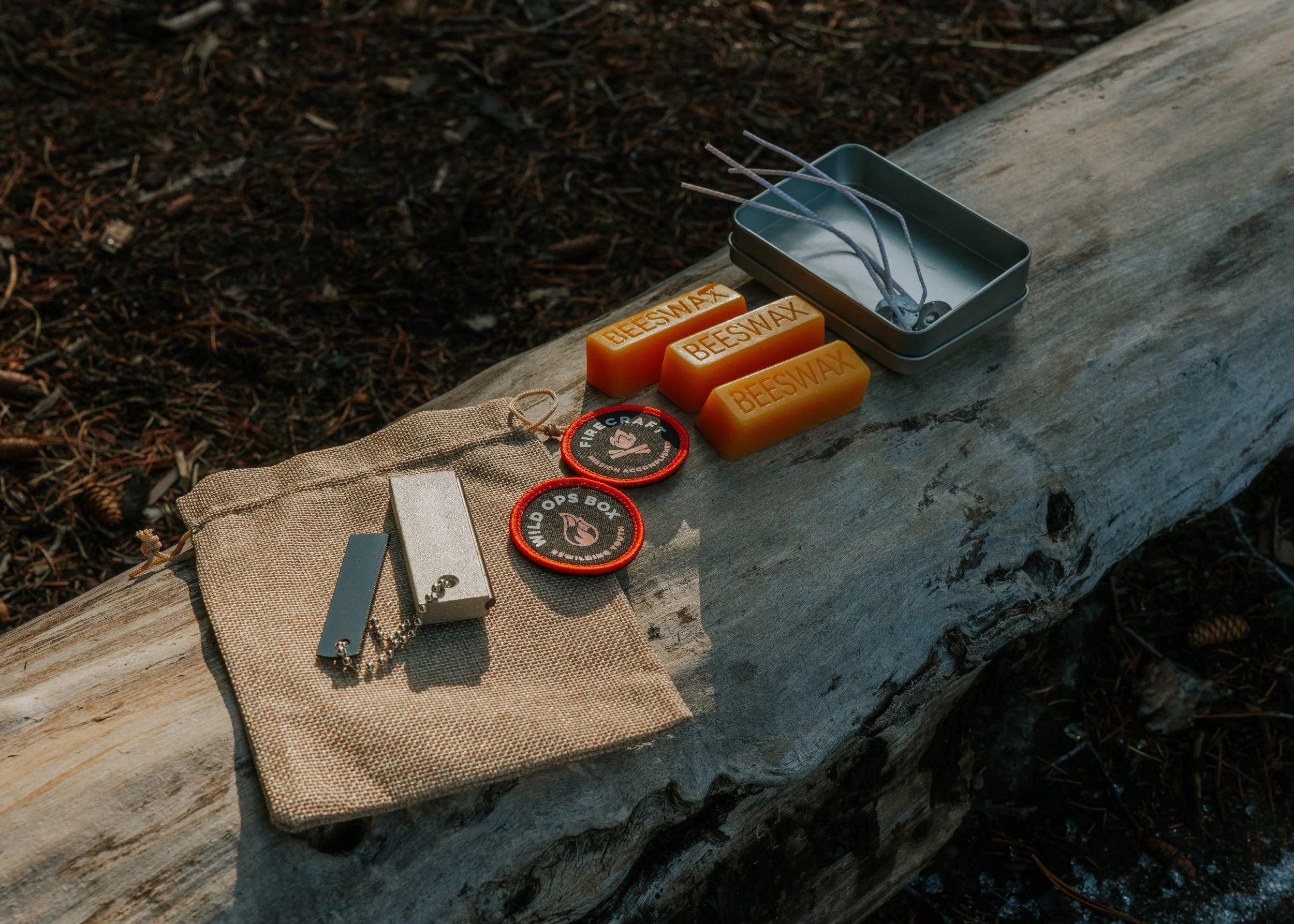 Outdoor scene with a wooden log holding various survival and outdoor gear, including beeswax soap, patches, a headlamp, and a storage box.
