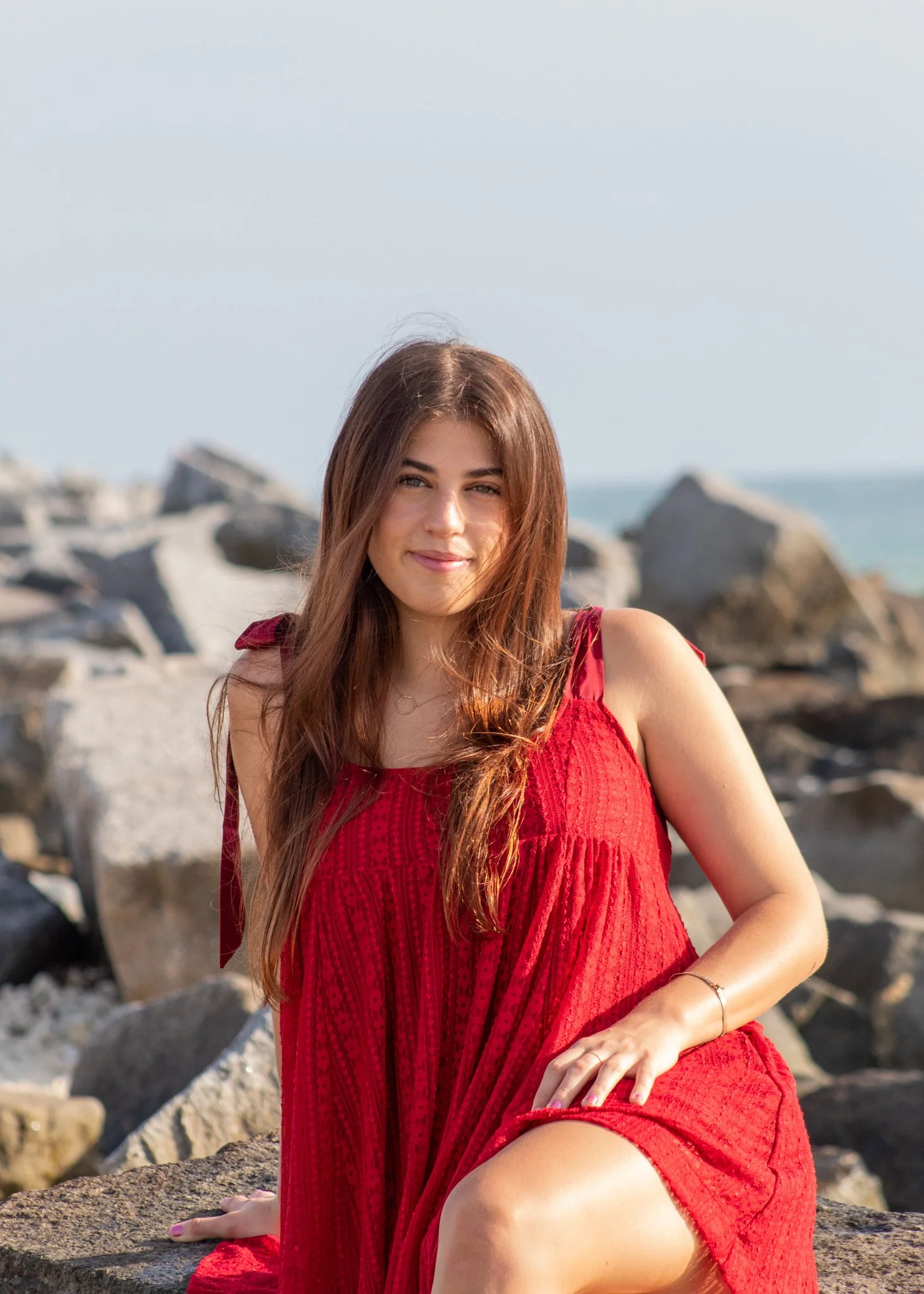 A woman with long brown hair sitting on rocks by the ocean, wearing a red dress.