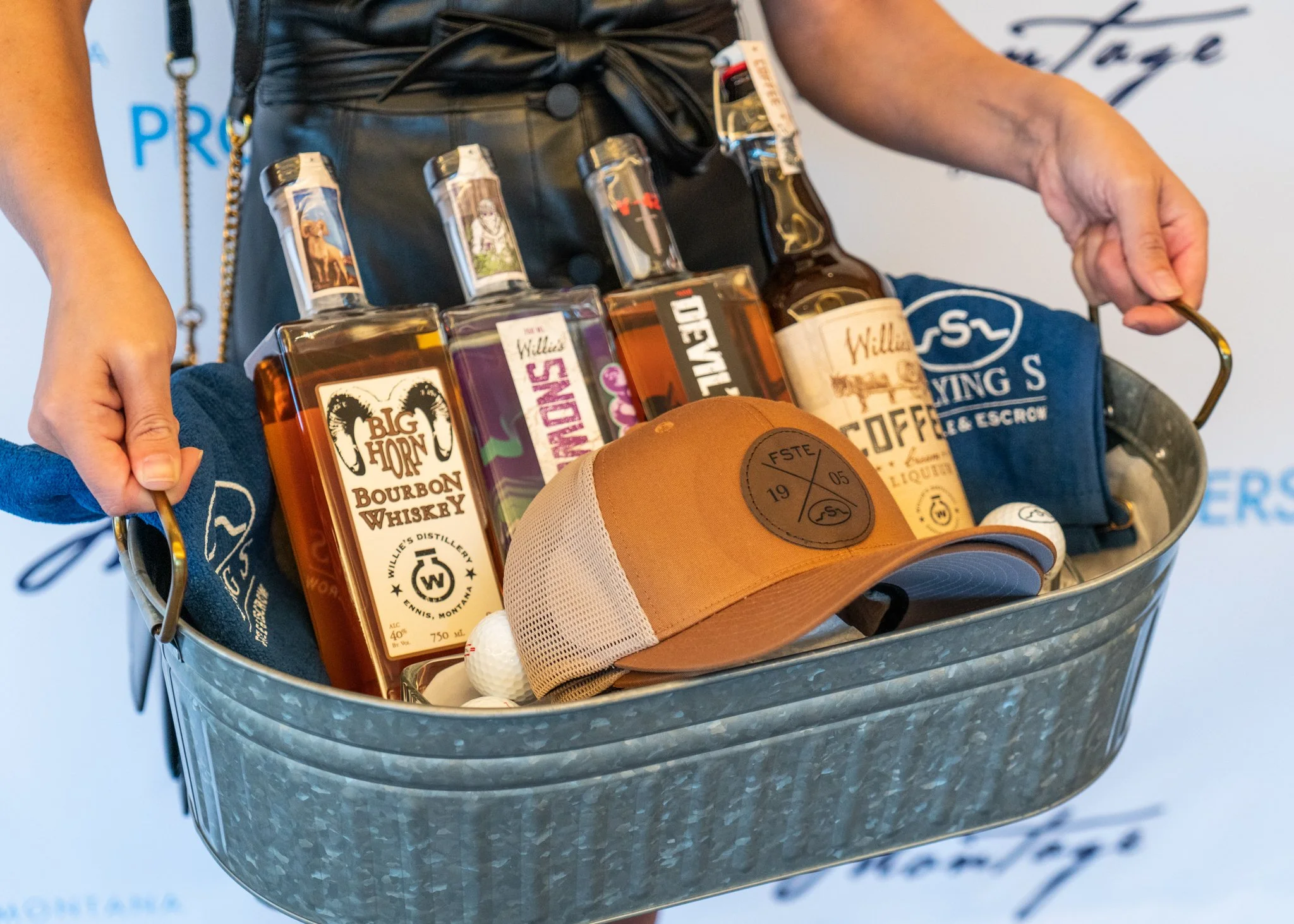 A person holding a metal bucket filled with several bottles of liquor, a baseball cap, and golf balls.