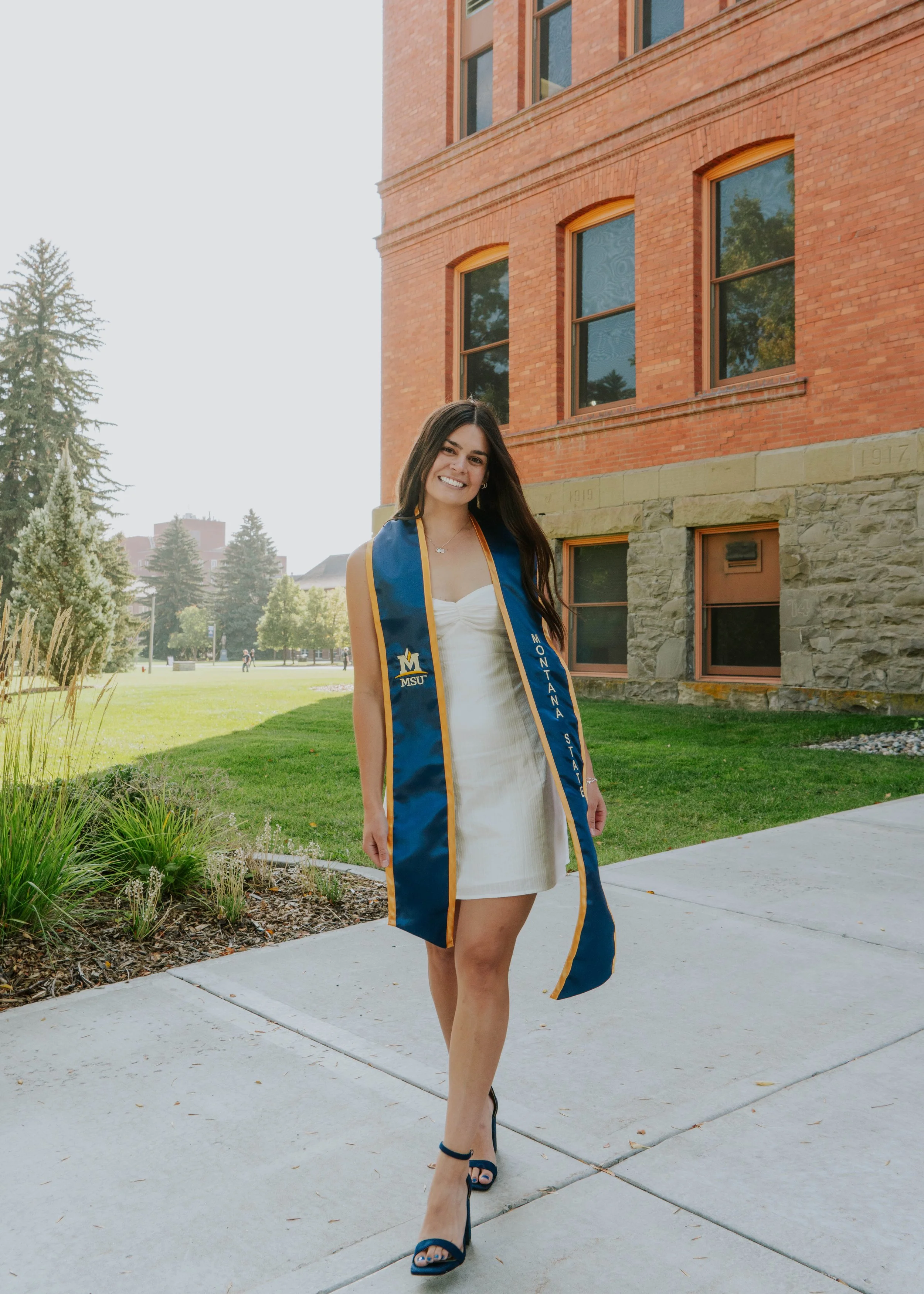 A young woman in a white dress and high heels smiling, wearing a blue graduation stole from Montana State University, standing outside in front of a brick building with large windows and green trees.