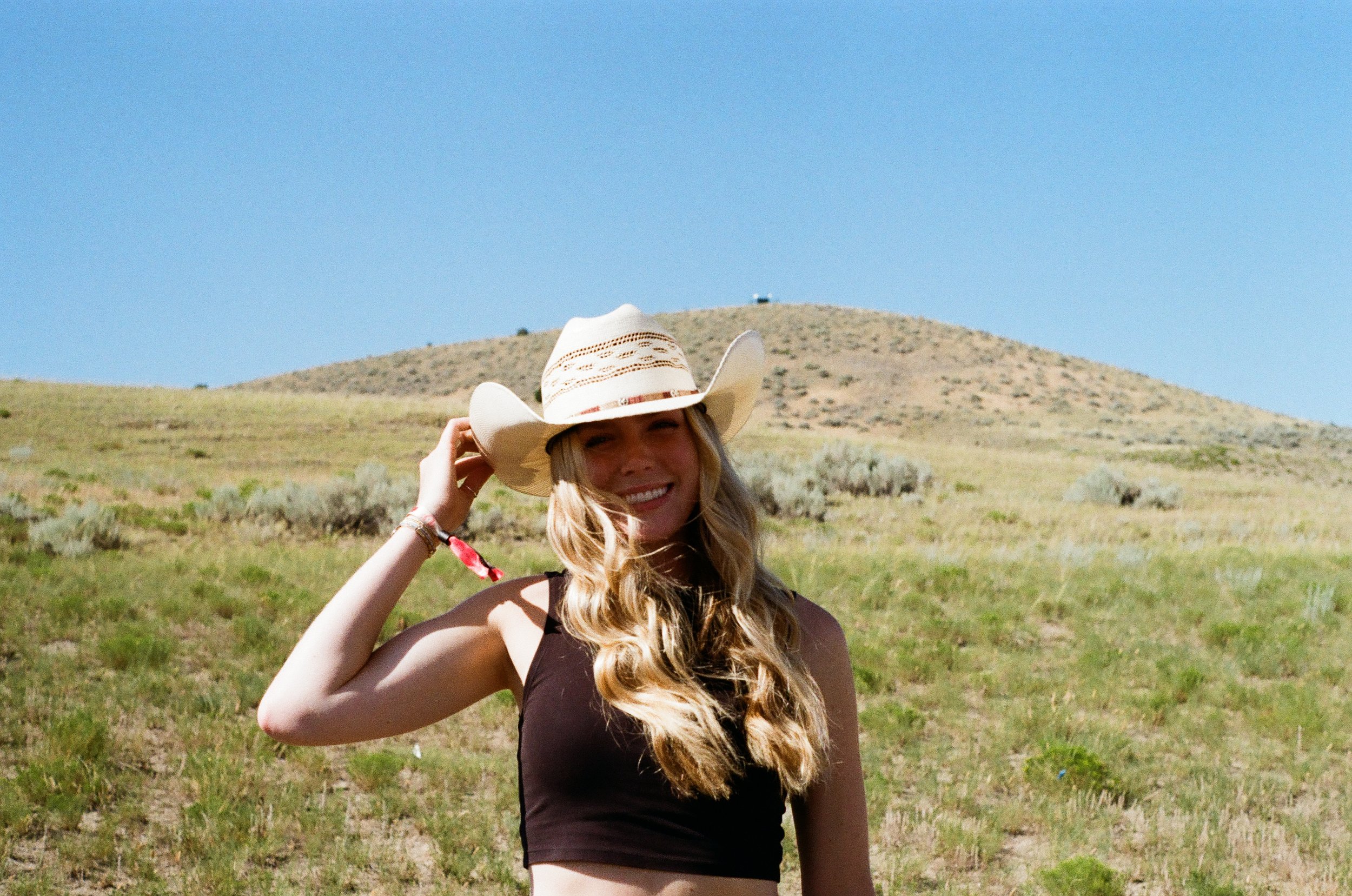 Young woman in a black crop top and wide-brimmed hat standing outdoors in a grassy field with a hill in the background under a clear blue sky.