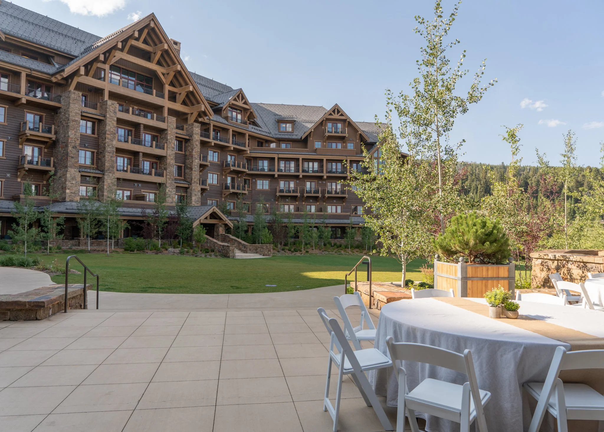 Outdoor patio with table and chairs in front of a grass lawn, trees, and a large multi-story wooden building with balconies and stone accents, set against a hillside and clear blue sky.