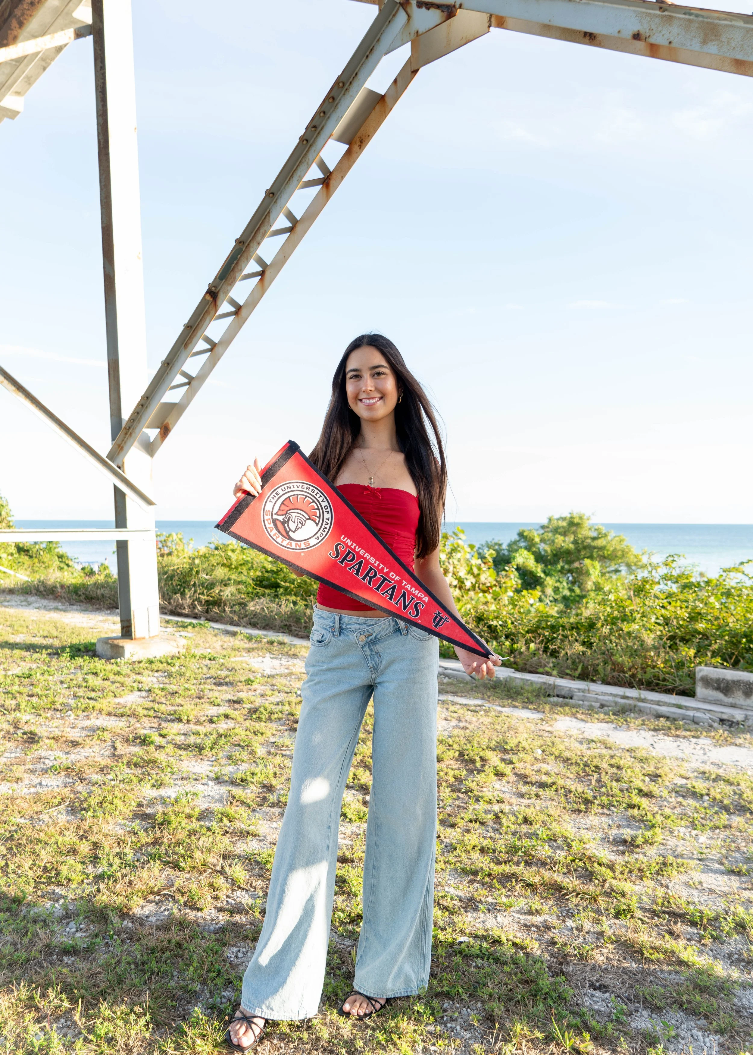 A young woman standing outdoors near the coast, holding a University of Tampa Spartans pennant, smiling, with a metal structure in the background and greenery around.