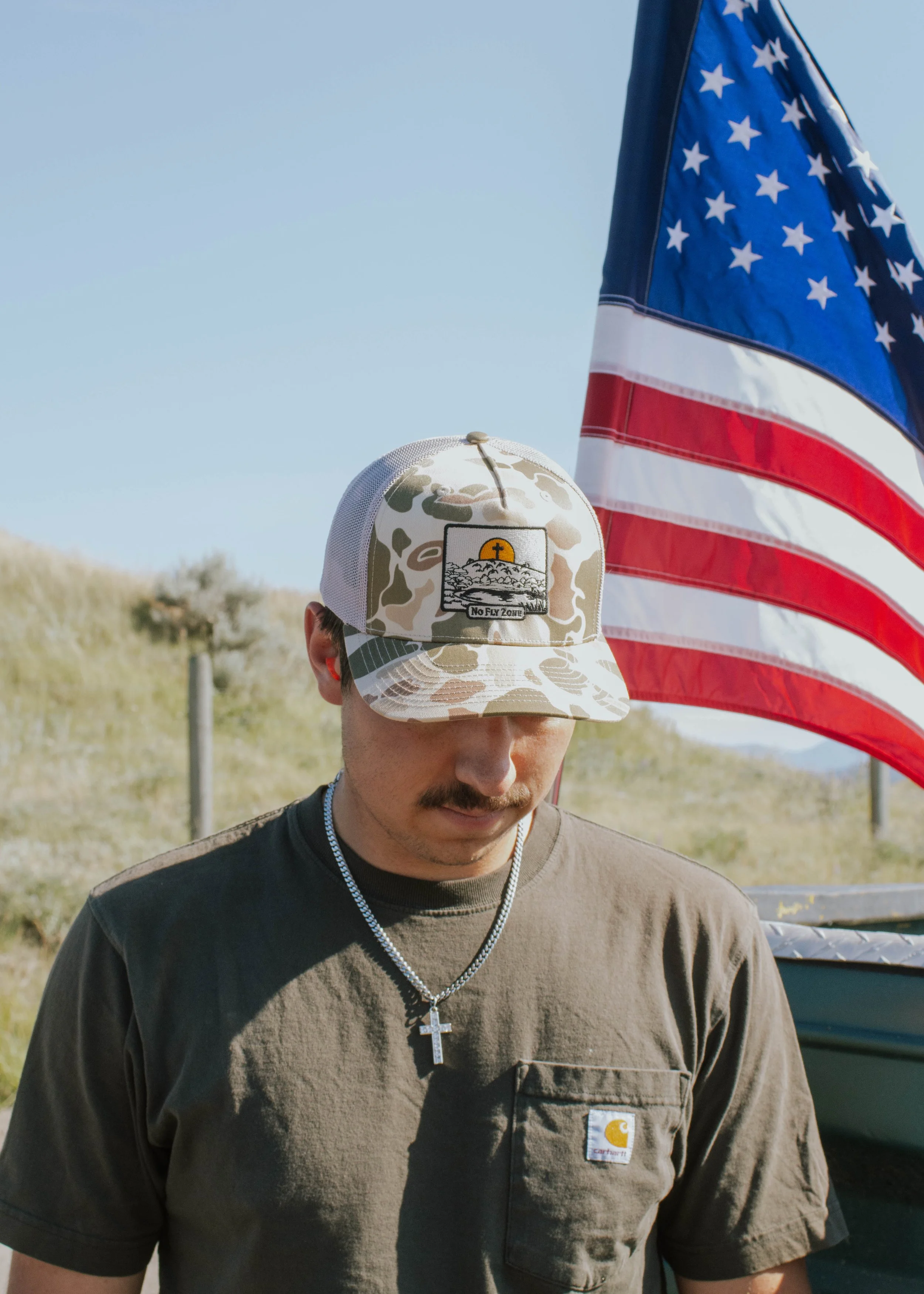 A man wearing a camouflage Carhartt cap with a patch that says 'No Fly Zone,' a brown Carhartt T-shirt, and a silver chain with a cross pendant. He appears to be looking down, standing outdoors with an American flag flying behind him and a grassy lan