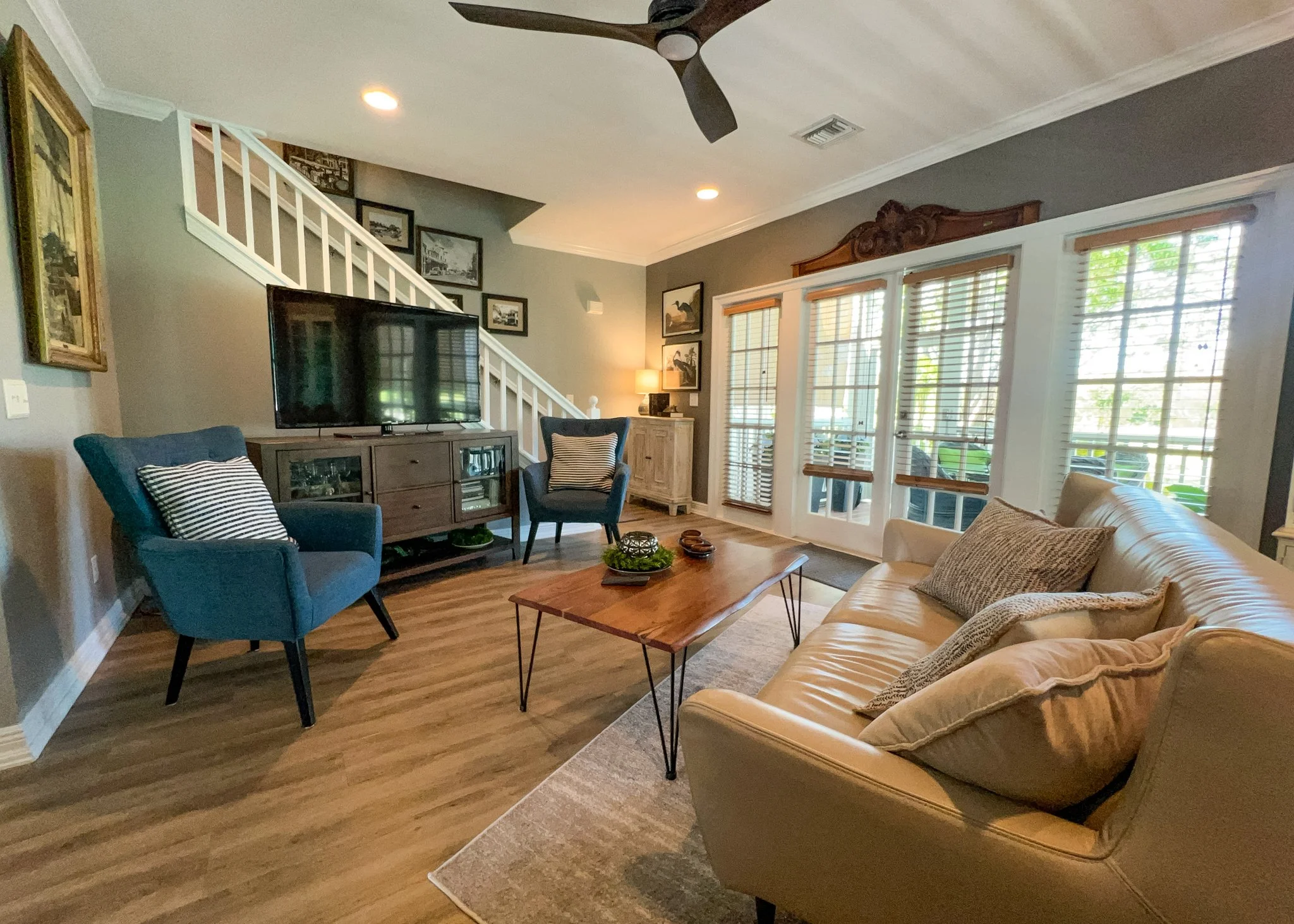 Living room with beige sofa, blue armchair, wooden coffee table, TV on stand, sliding glass door with blinds, staircase, and framed pictures on the wall.