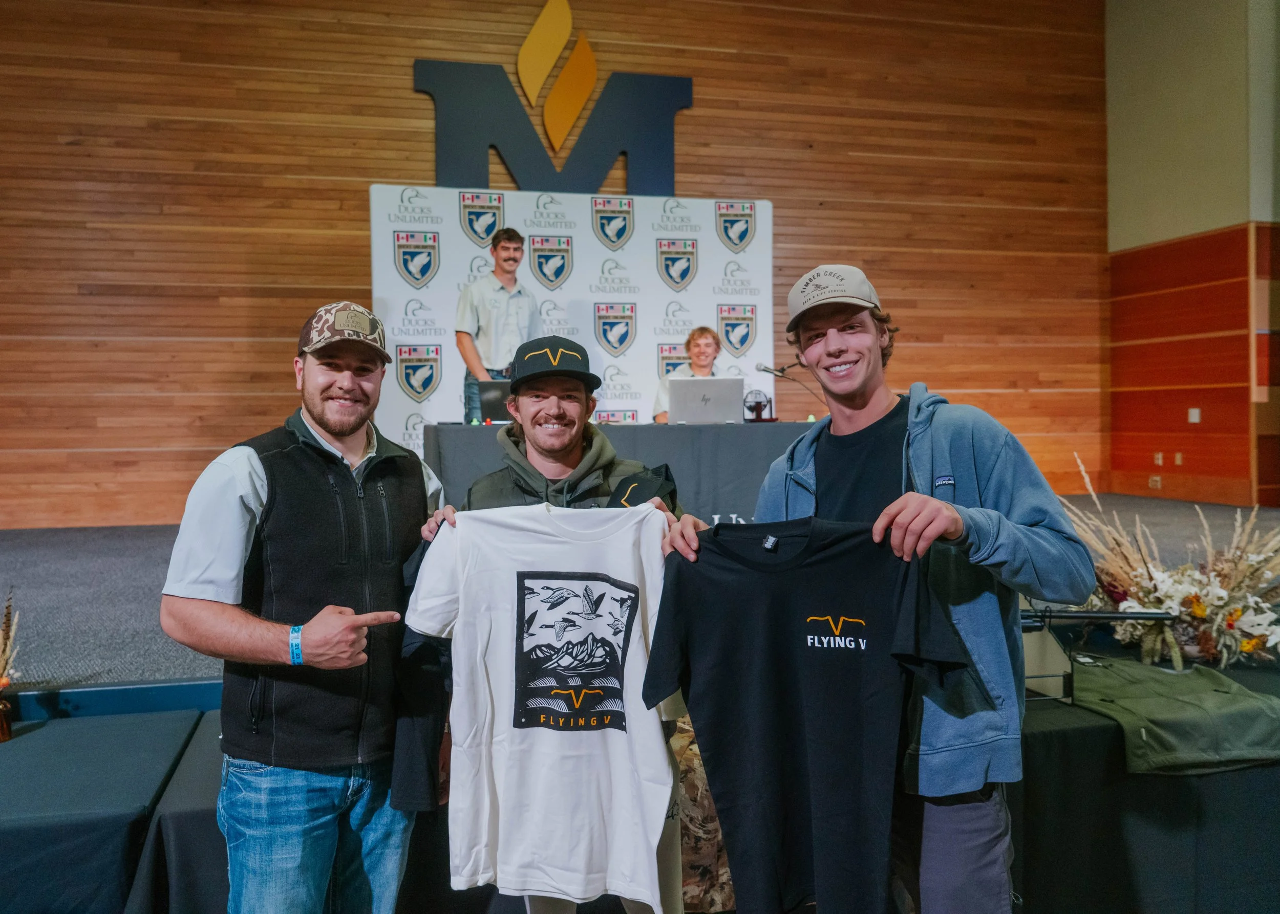 Three young men smiling and holding up T-shirts at an indoor event, with a media table and two men seated at a table in the background. The setting features a large wooden wall and a logo on the backdrop.