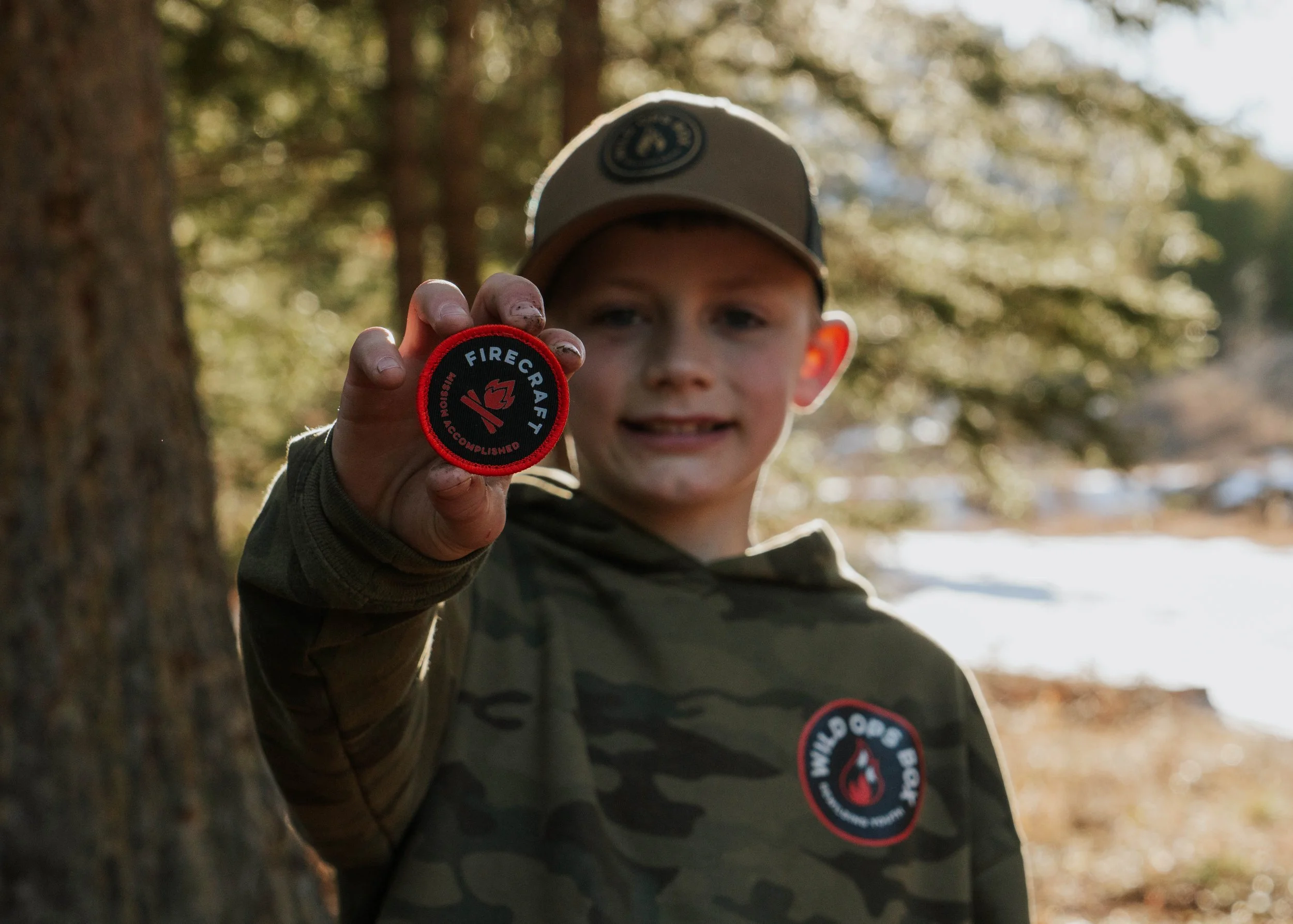 A young boy holding a Firecraft patch in an outdoor wooded area with trees and some snow in the background.