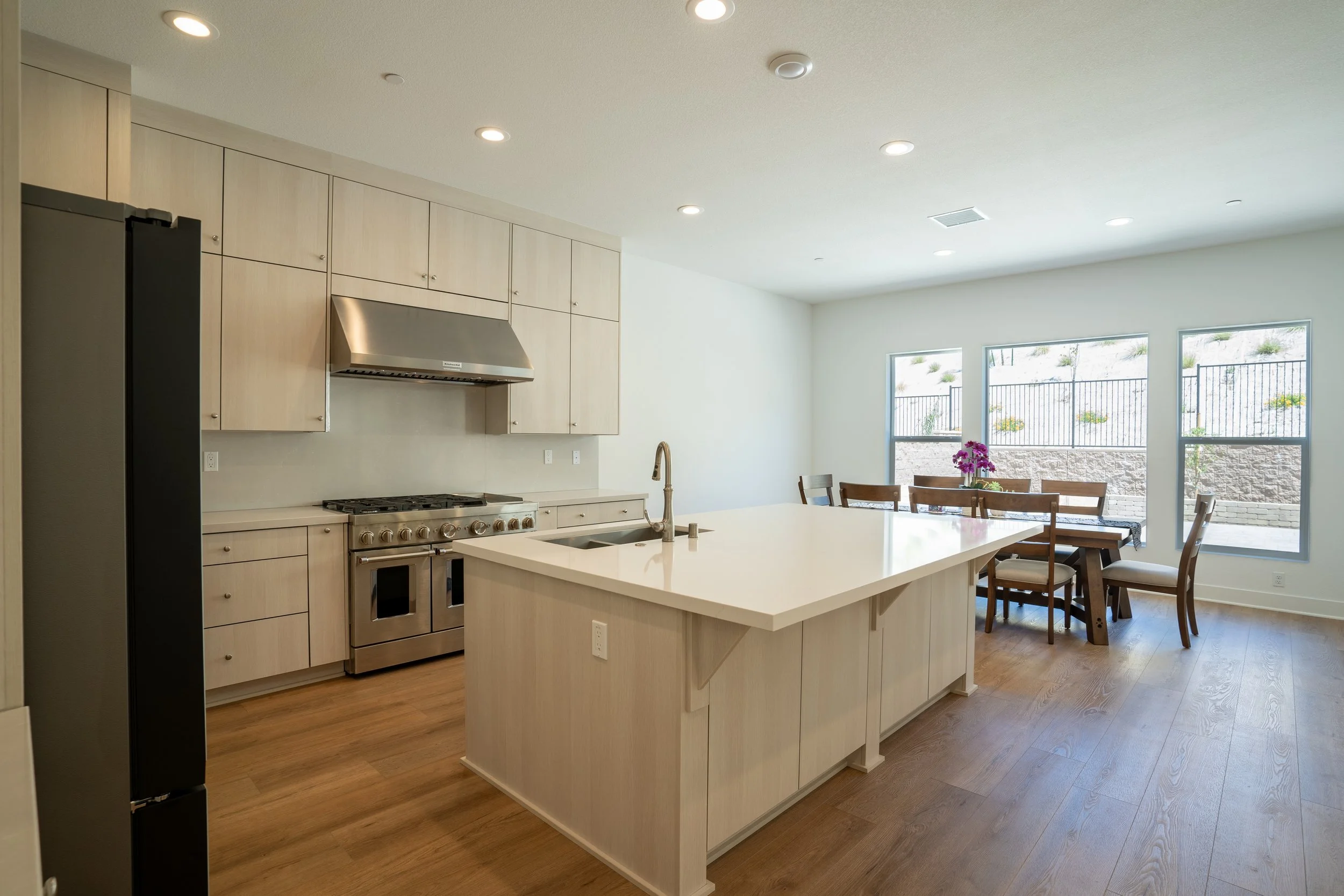 Modern kitchen with light wood cabinets and an island, stainless steel stove, and a dining area with a table, chairs, and windows with a view of an outdoor yard with a fence and plants.