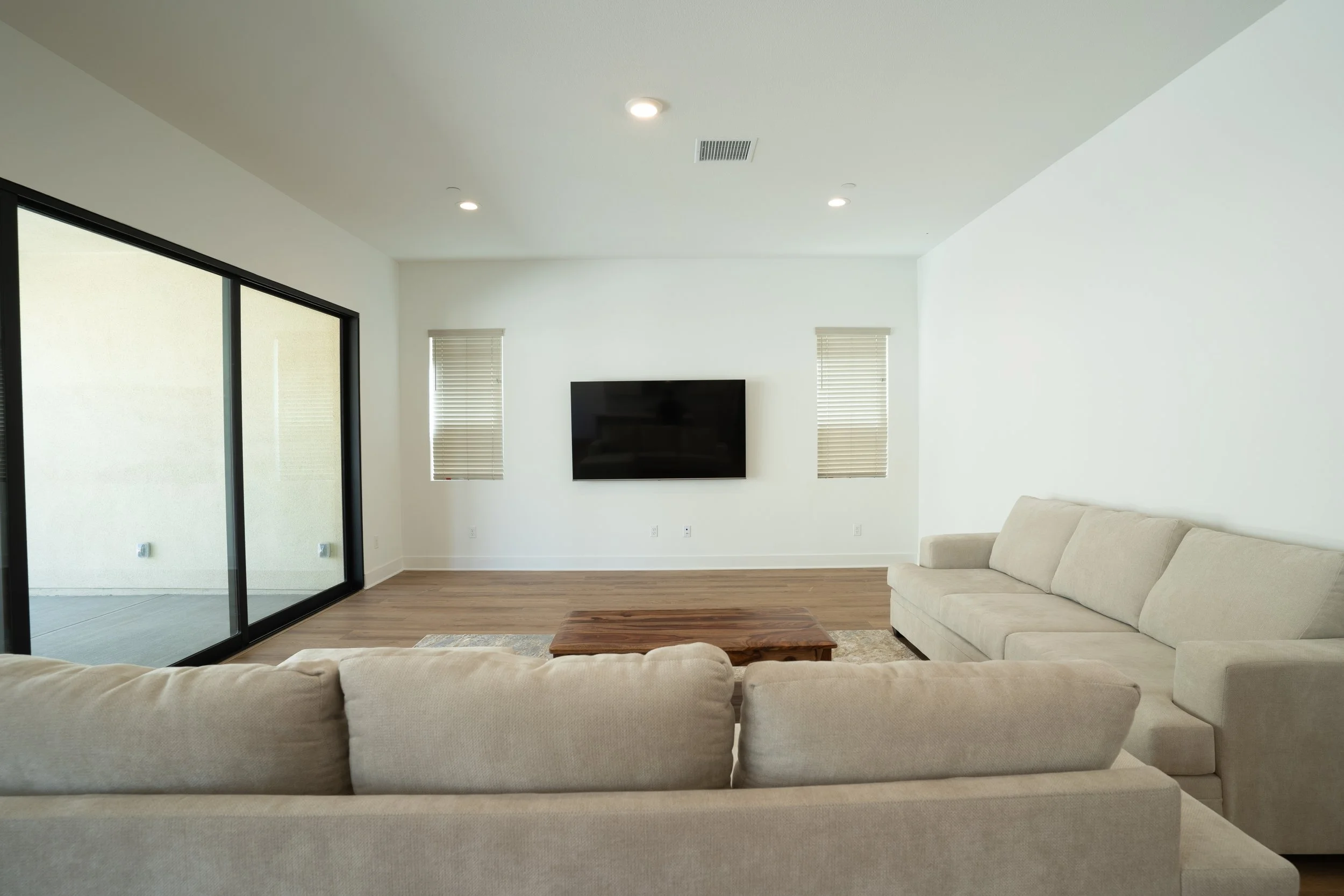 Living room with beige sofas, a wooden coffee table, a wall-mounted TV, two small windows with blinds, a sliding glass door, and a hardwood floor.