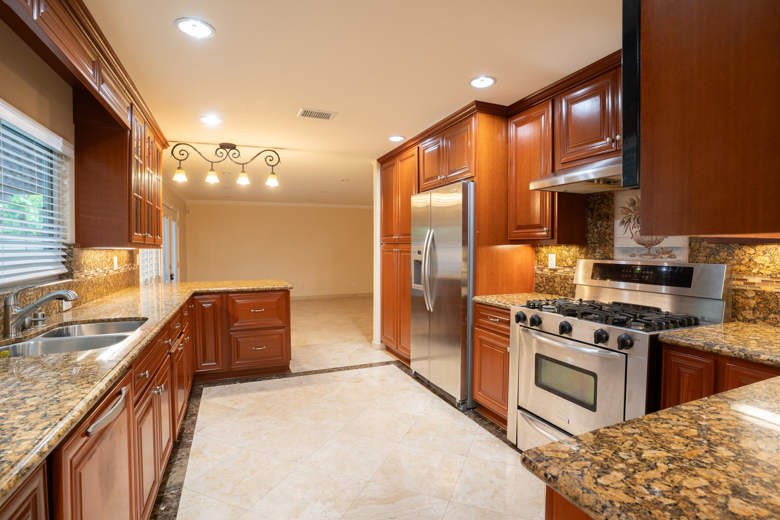 Clean kitchen with wooden cabinets, granite countertops, stainless steel refrigerator and stove, tiled floor, and a window with blinds.