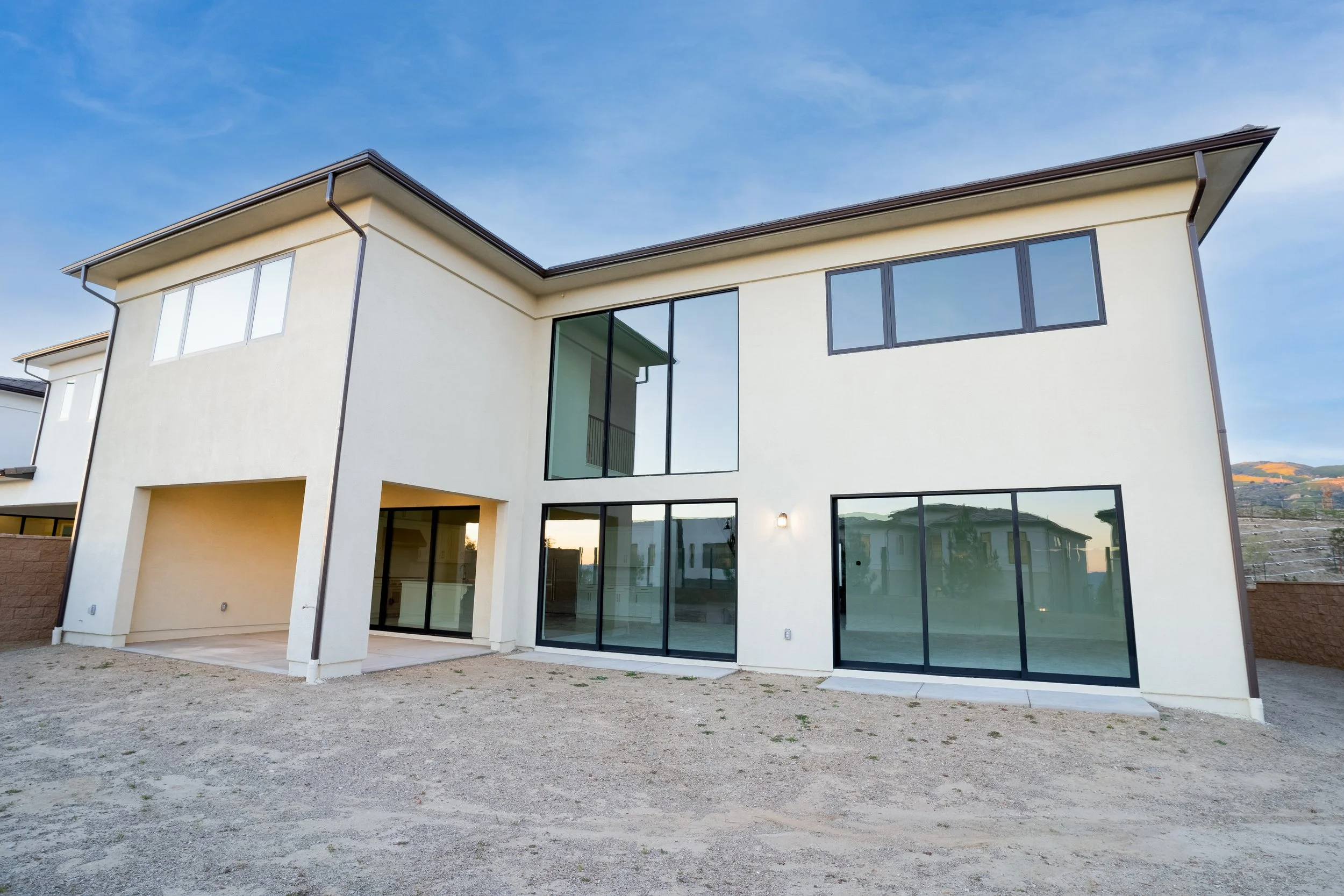 A modern two-story house with large glass windows and a beige exterior, situated in a semi-rural area with a dirt yard and a mountain in the background under a blue sky.