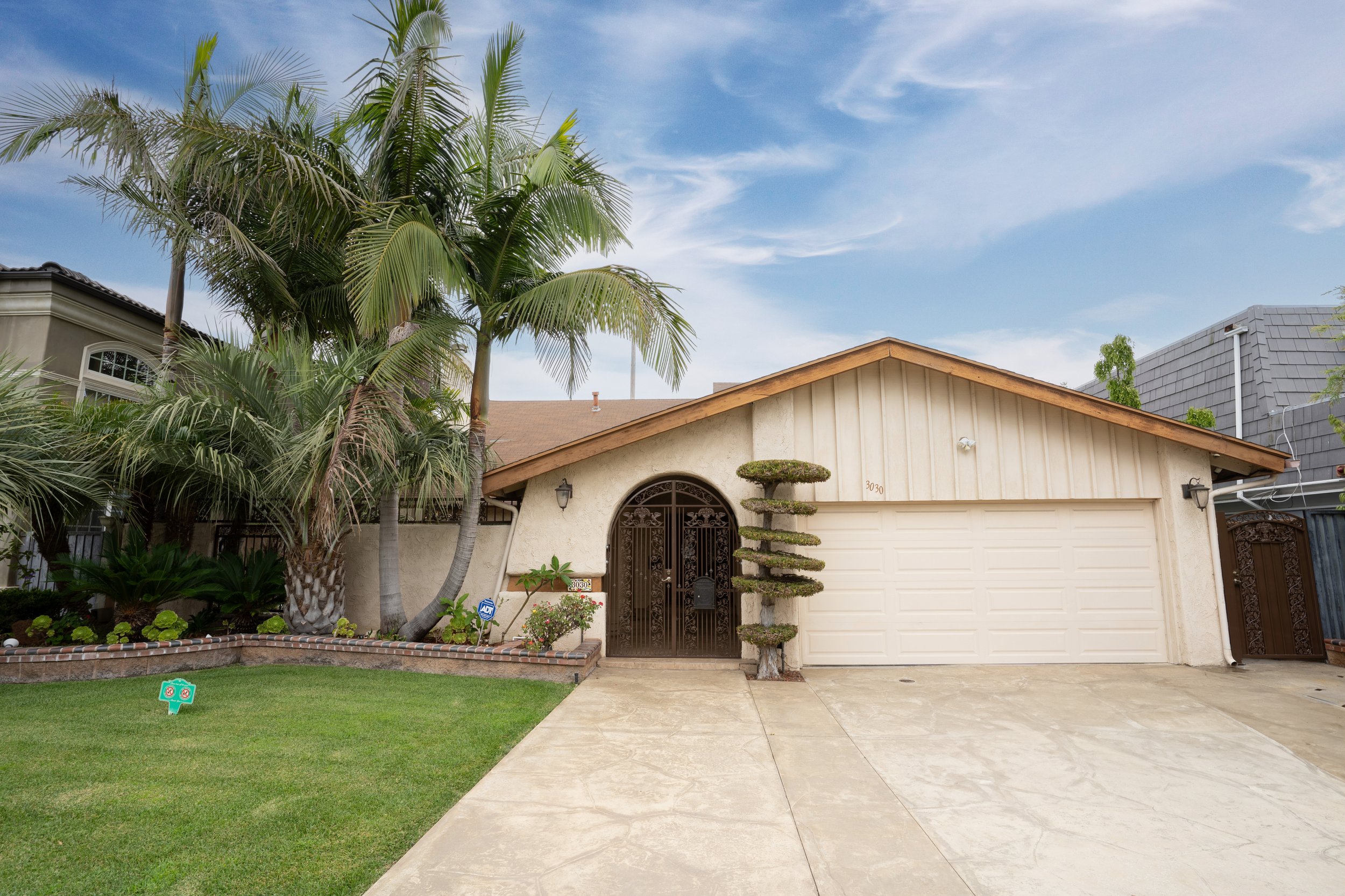 A house with a white garage door, dark front door with an ornate iron gate, surrounded by various tropical plants including tall palm trees, a multi-tiered trimmed bush, and a small floral garden. The sky is partly cloudy and the driveway is concrete.