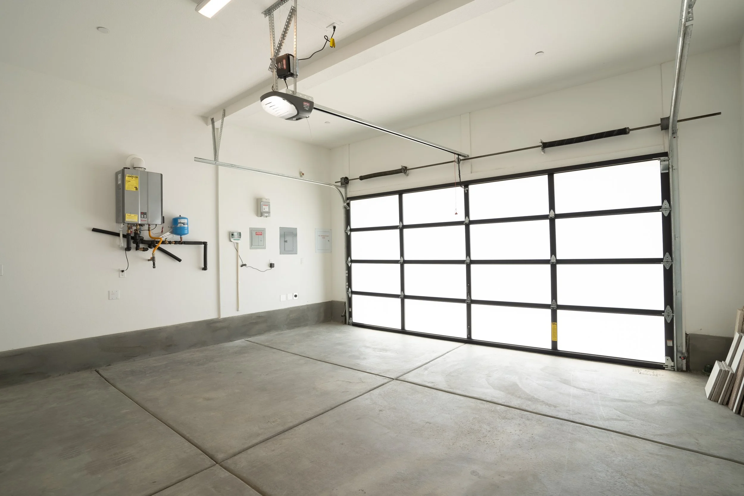 Empty garage with a large closed black metal garage door, concrete floor, white walls, and electrical equipment on the wall.