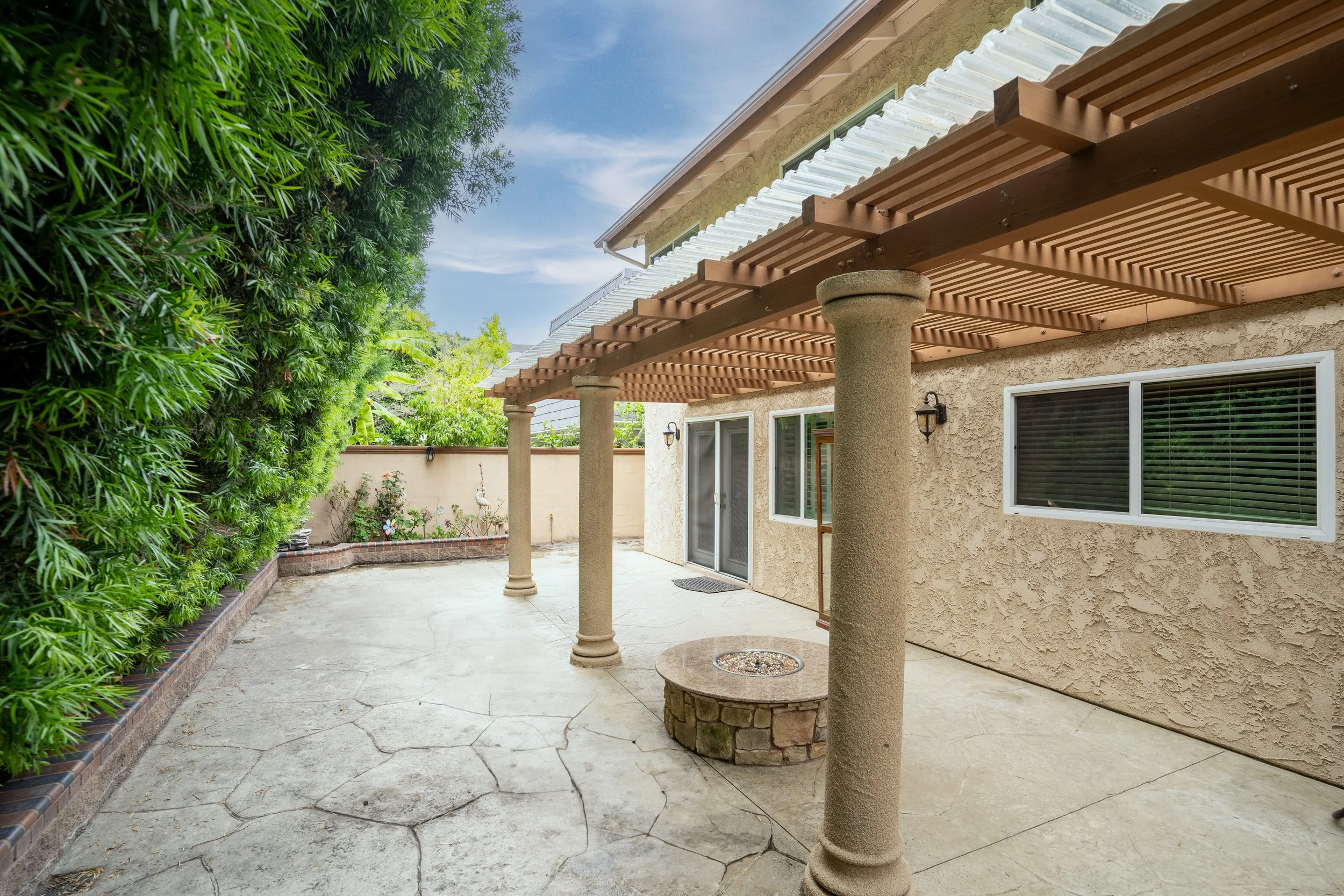 Backyard patio with decorative columns supporting a wooden pergola, concrete flooring, a built-in fire pit, and a beige stucco house wall with windows and outdoor lights, surrounded by green trees and plants.