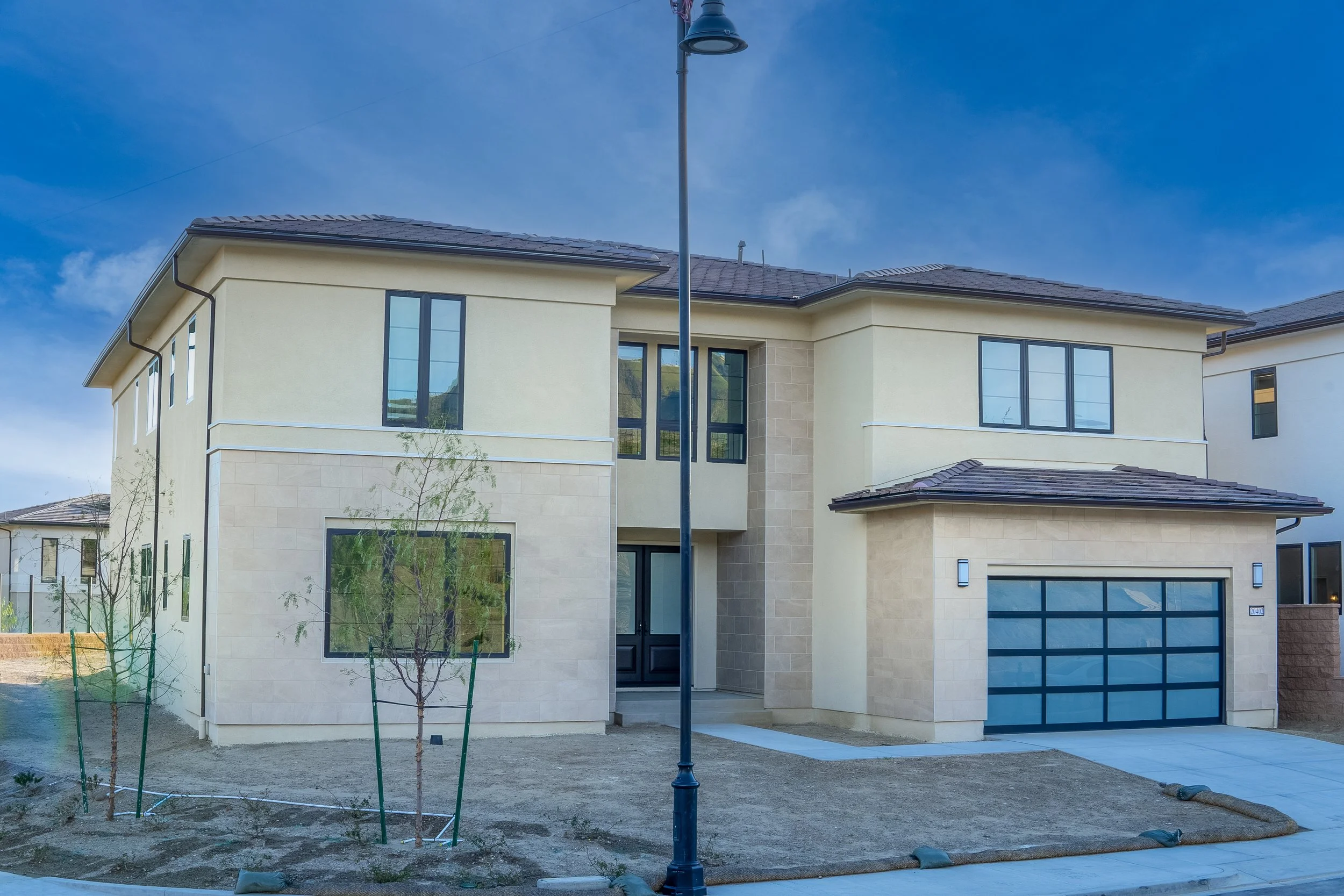 Modern two-story house with light-colored exterior walls, a dark gray garage door, and a small front yard with young trees, under a partly cloudy sky.