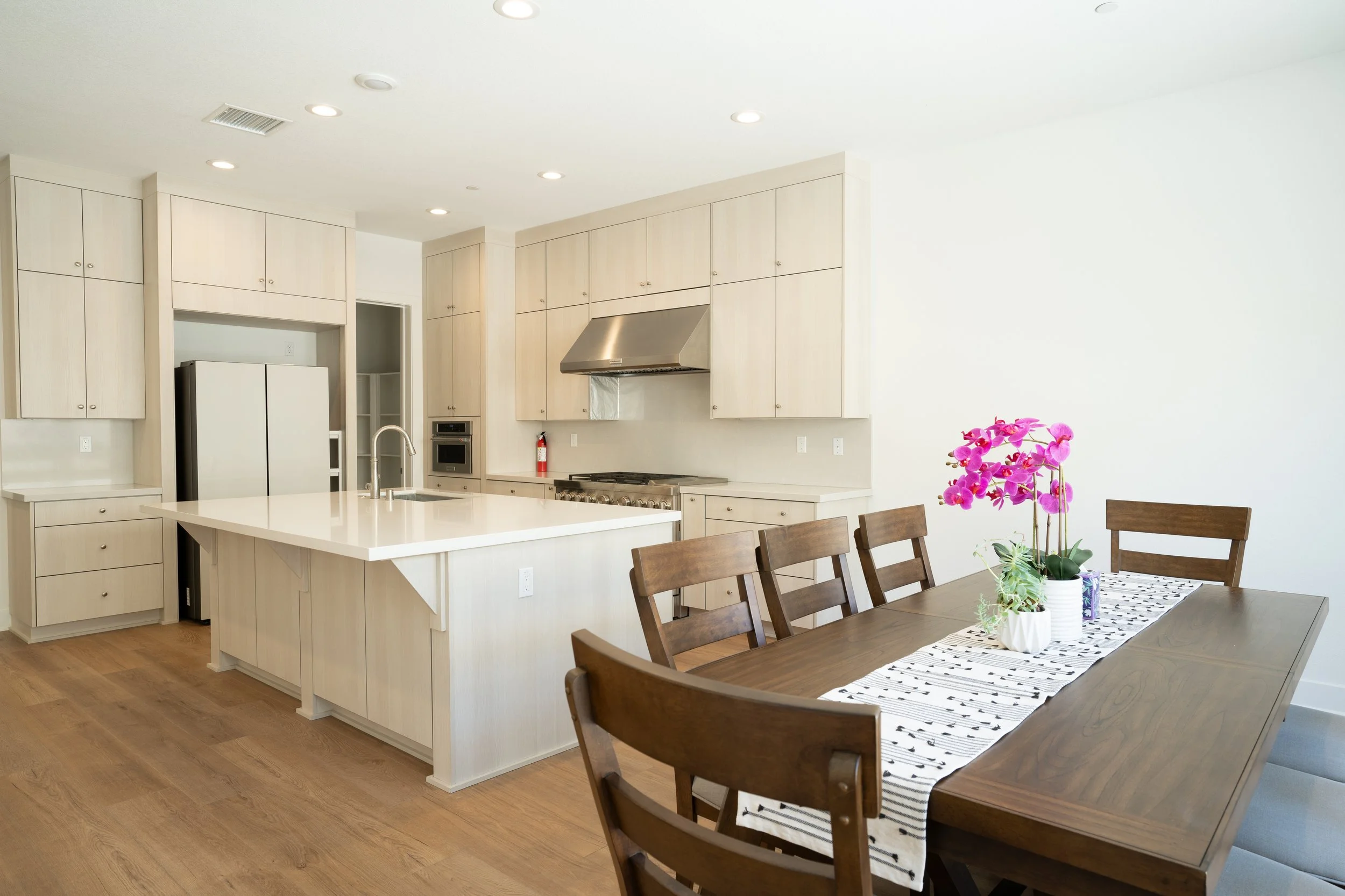 Modern kitchen with beige cabinets, white island, stainless steel range hood, and a dining table with wood chairs and pink orchids in a white pot.