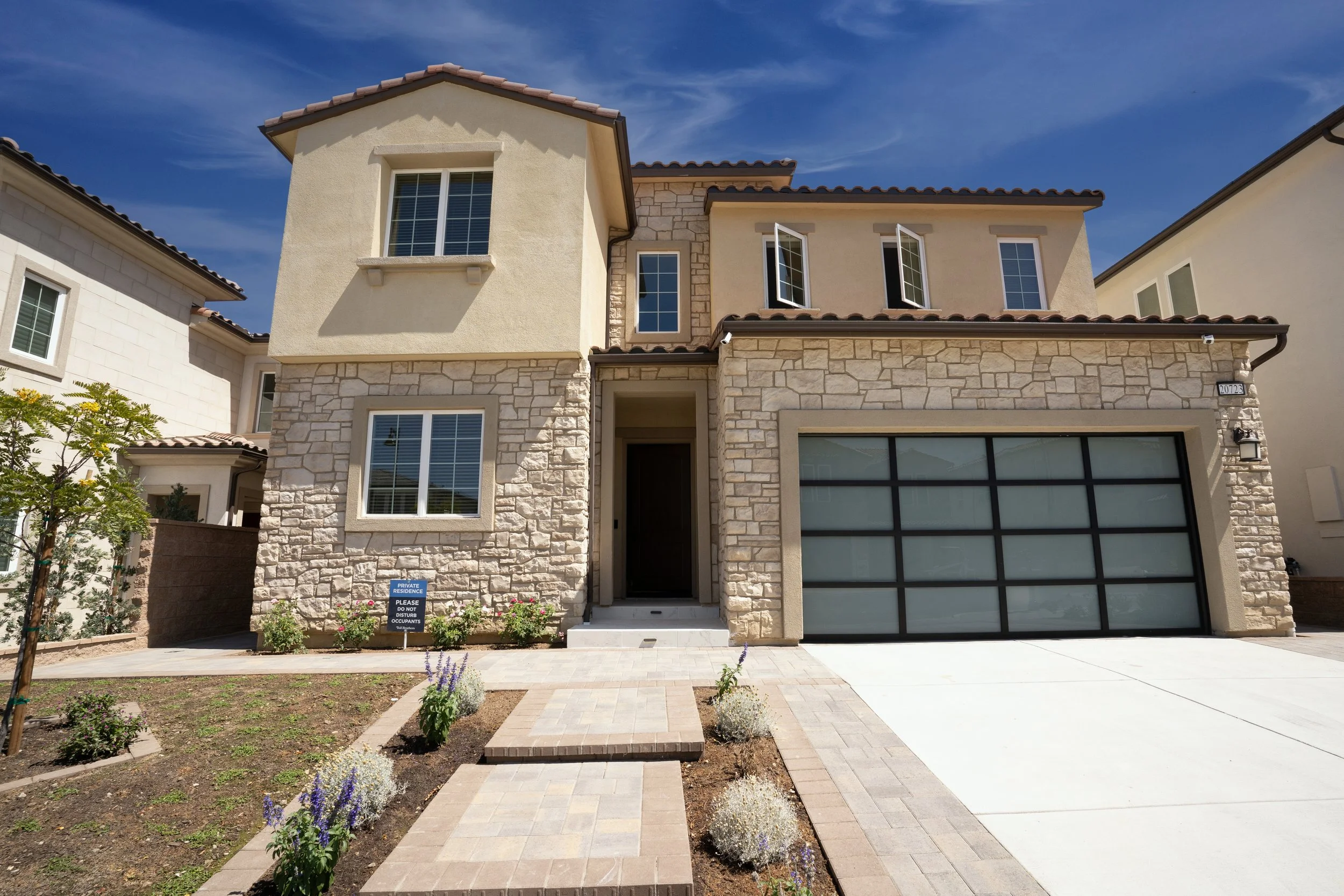 Front view of a modern two-story house with stone and stucco exterior, a garage with a glass-paneled door, and a paved walkway with landscaping