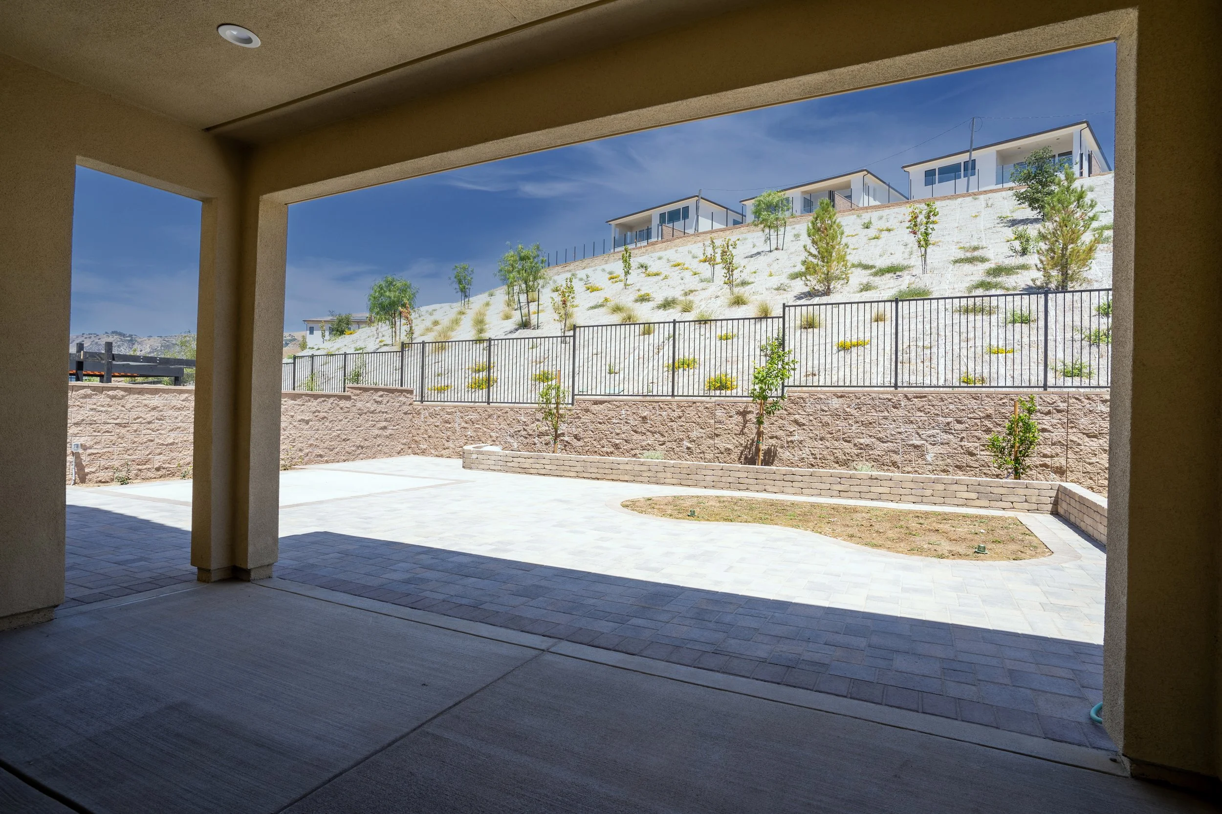 View from a covered patio looking out over a landscaped yard with small trees, a stone retaining wall, and a hill with more trees and modern houses on top, under a blue sky.
