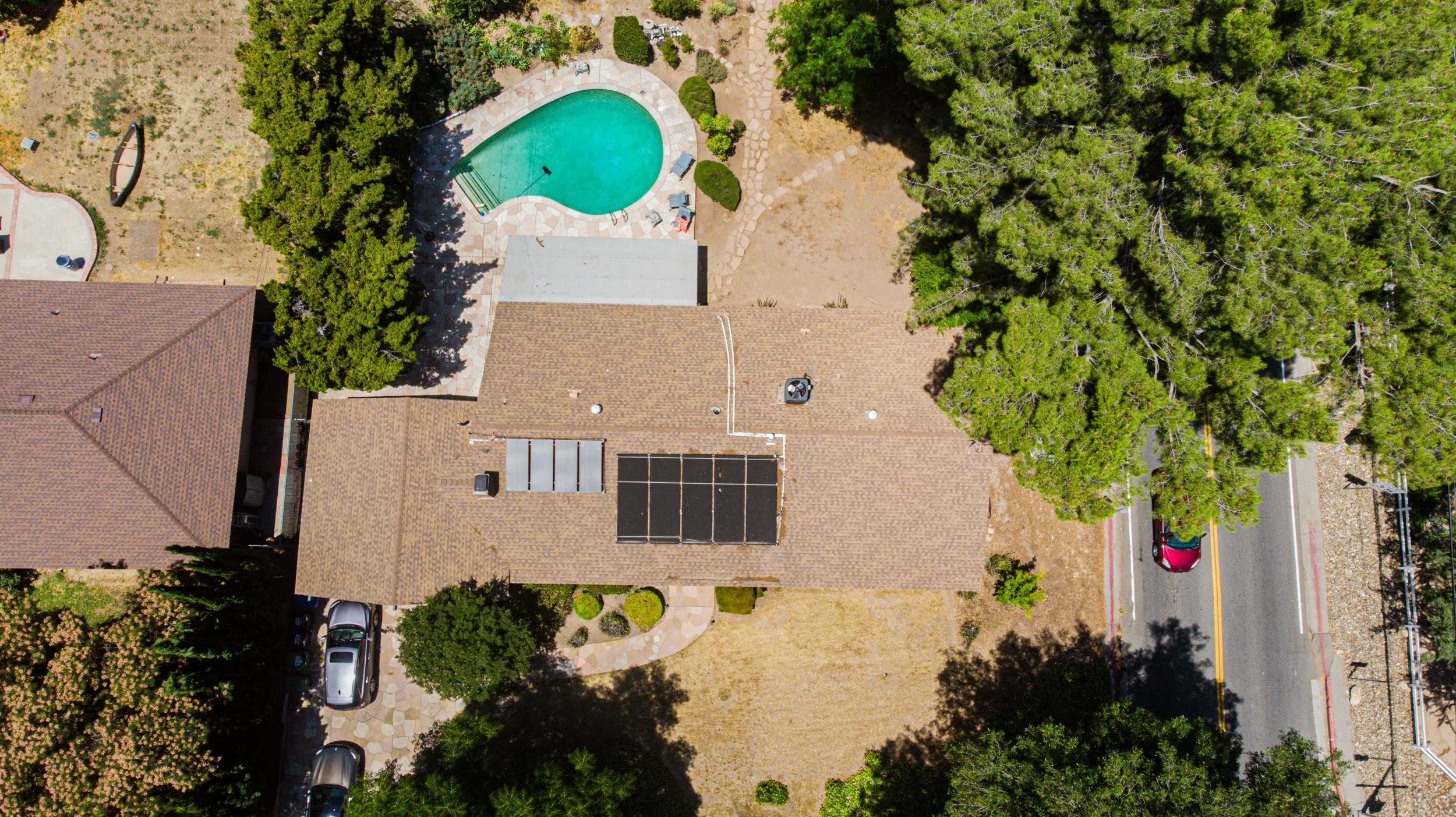 An aerial view of a residential backyard with a swimming pool, surrounding trees, a brick house, a driveway with parked cars, and a street with a red car driving by.