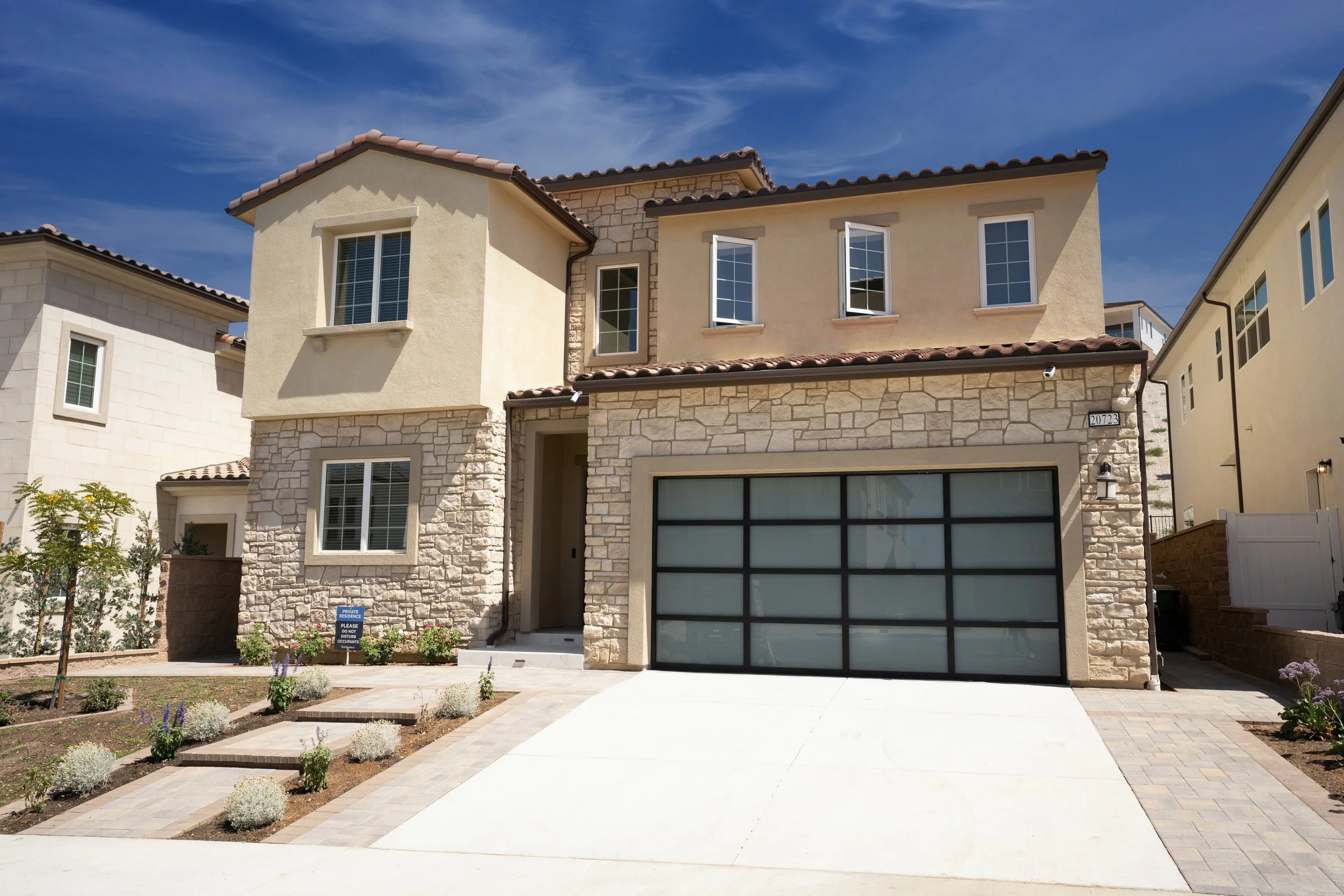 Modern two-story house with stone and stucco exterior, rectangular windows, a dark garage door, and a small front yard with plants and a concrete driveway.