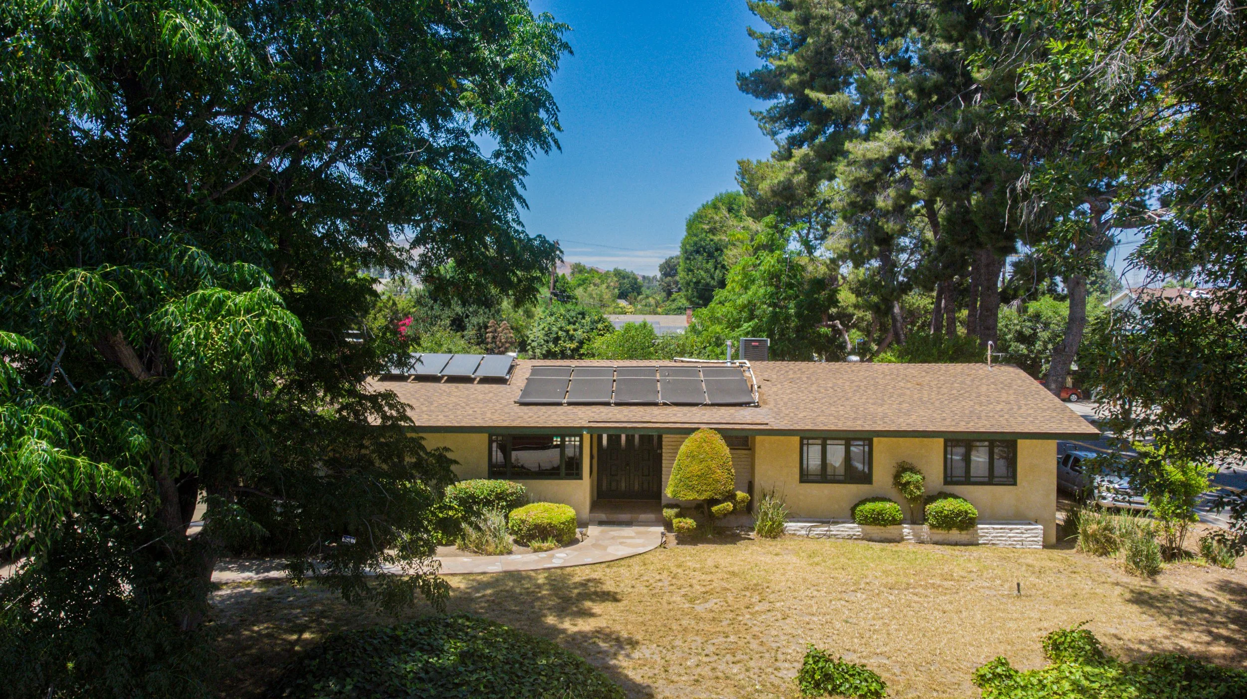 A beige single-story house with windows on both sides, a front door, landscaped bushes, and trees, with solar panels on the roof, under a clear blue sky.