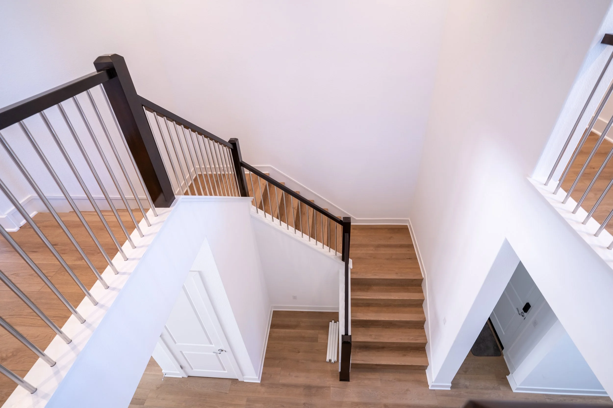 Interior view of a modern staircase with wooden steps and a dark wood handrail, viewed from above, showing white walls and a hardwood floor.