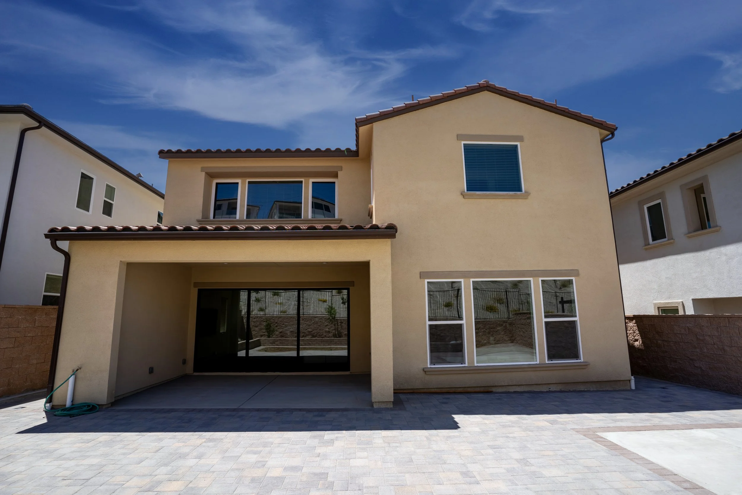 A two-story beige house with a tiled roof, large windows, and a covered carport. There are neighboring homes on either side, and the sky is partly cloudy.