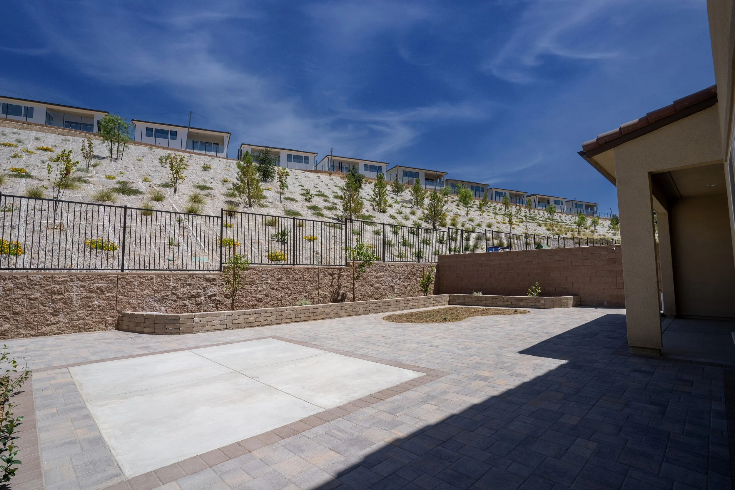 A spacious backyard patio with a paved surface, a small circular garden, and a retaining wall with a fence. A hillside with decorative plants and trees rises behind the fence, and a row of modern small houses or apartments is visible at the top under