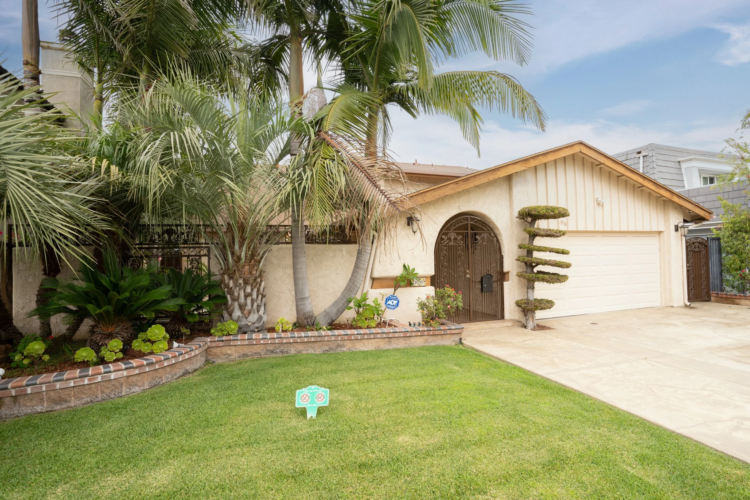 Front yard of a house with a well-maintained green lawn, tropical plants, and palm trees. The house has a beige exterior with a brown arched front door, a white garage door, and a decorative topiary.