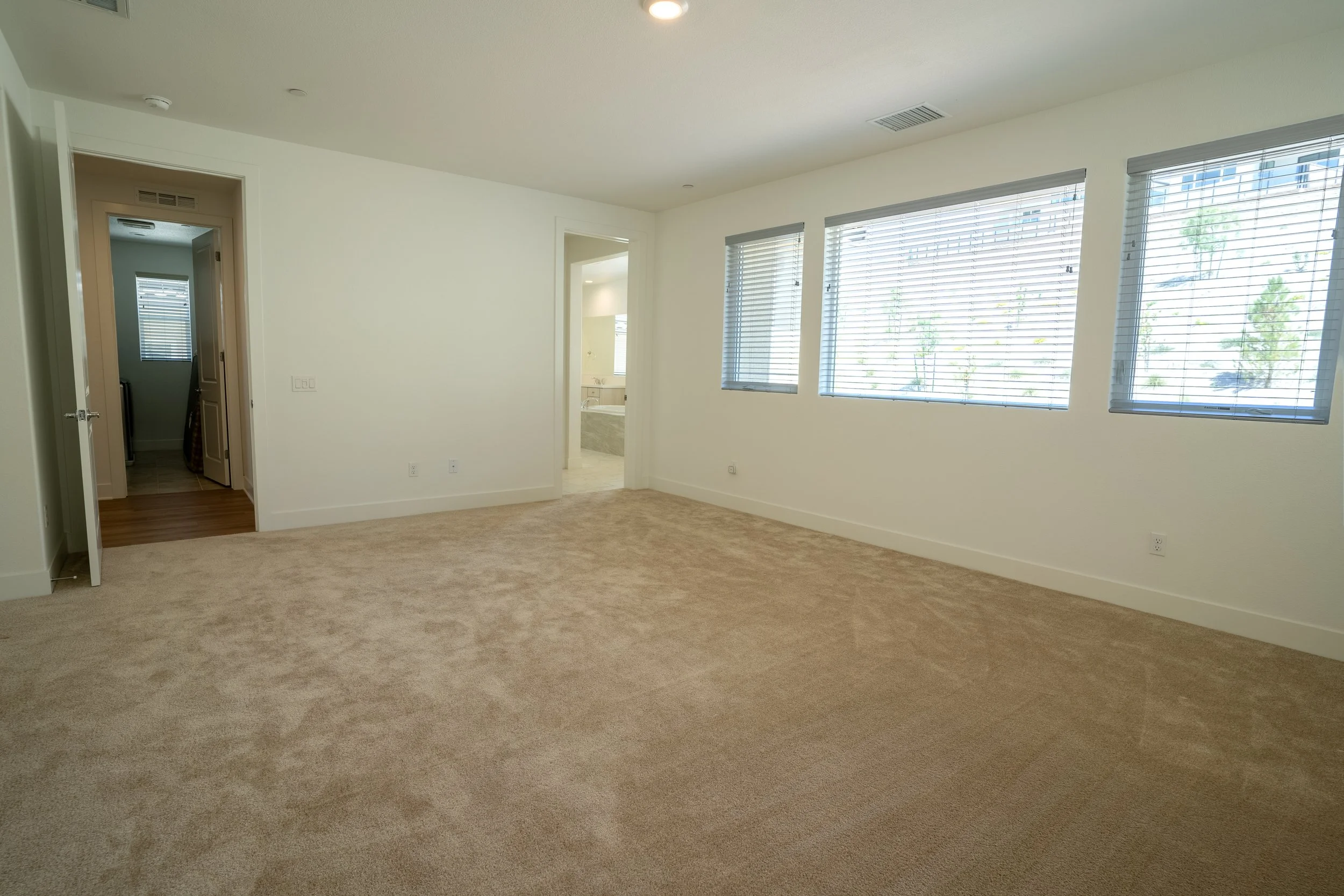 Empty living room with large windows, beige carpet, white walls, and an open doorway leading to a bathroom.