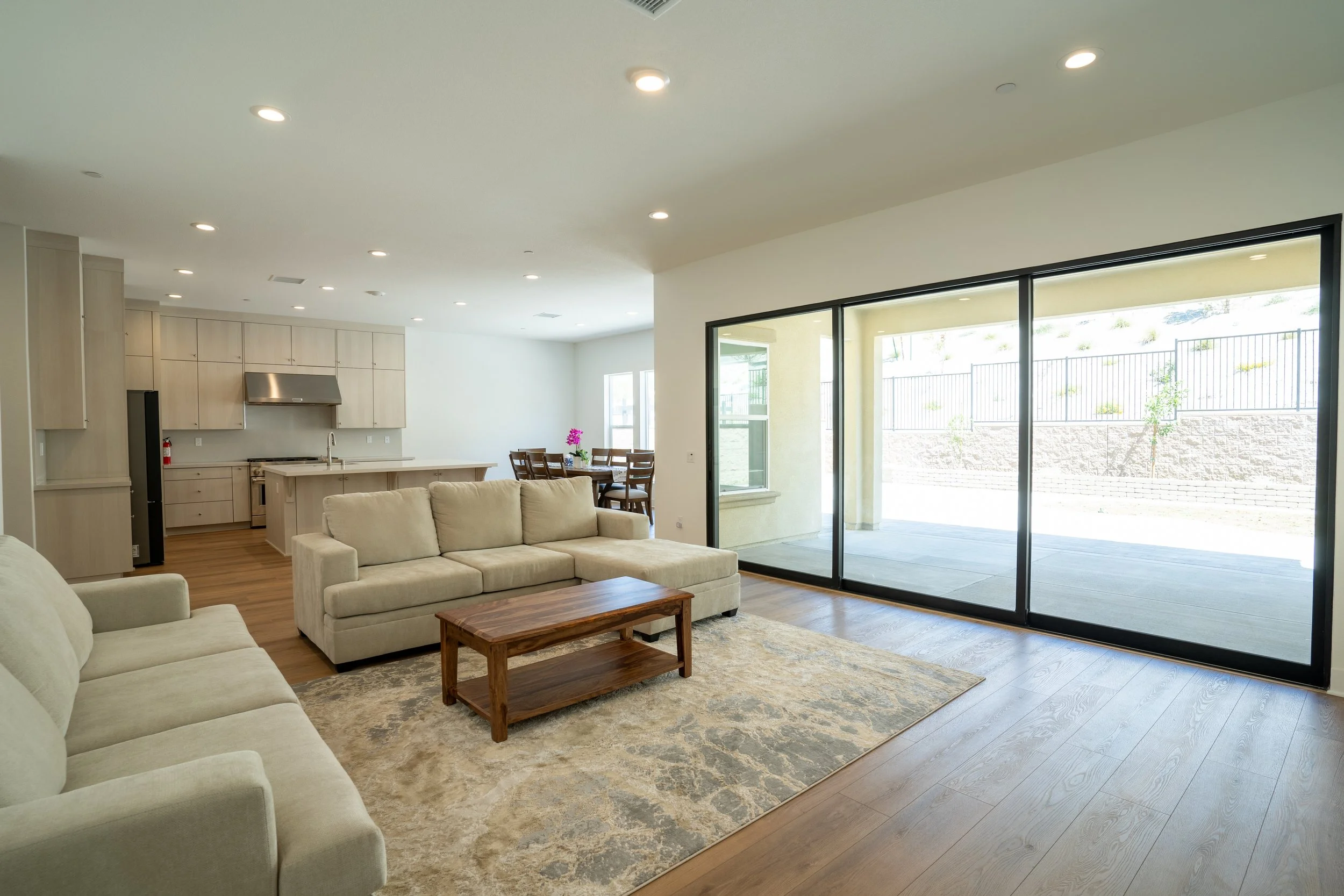 Open-concept living room and kitchen area with sliding glass doors leading to patio, beige sofas, wooden coffee table, dining table with chairs, and neutral tones.
