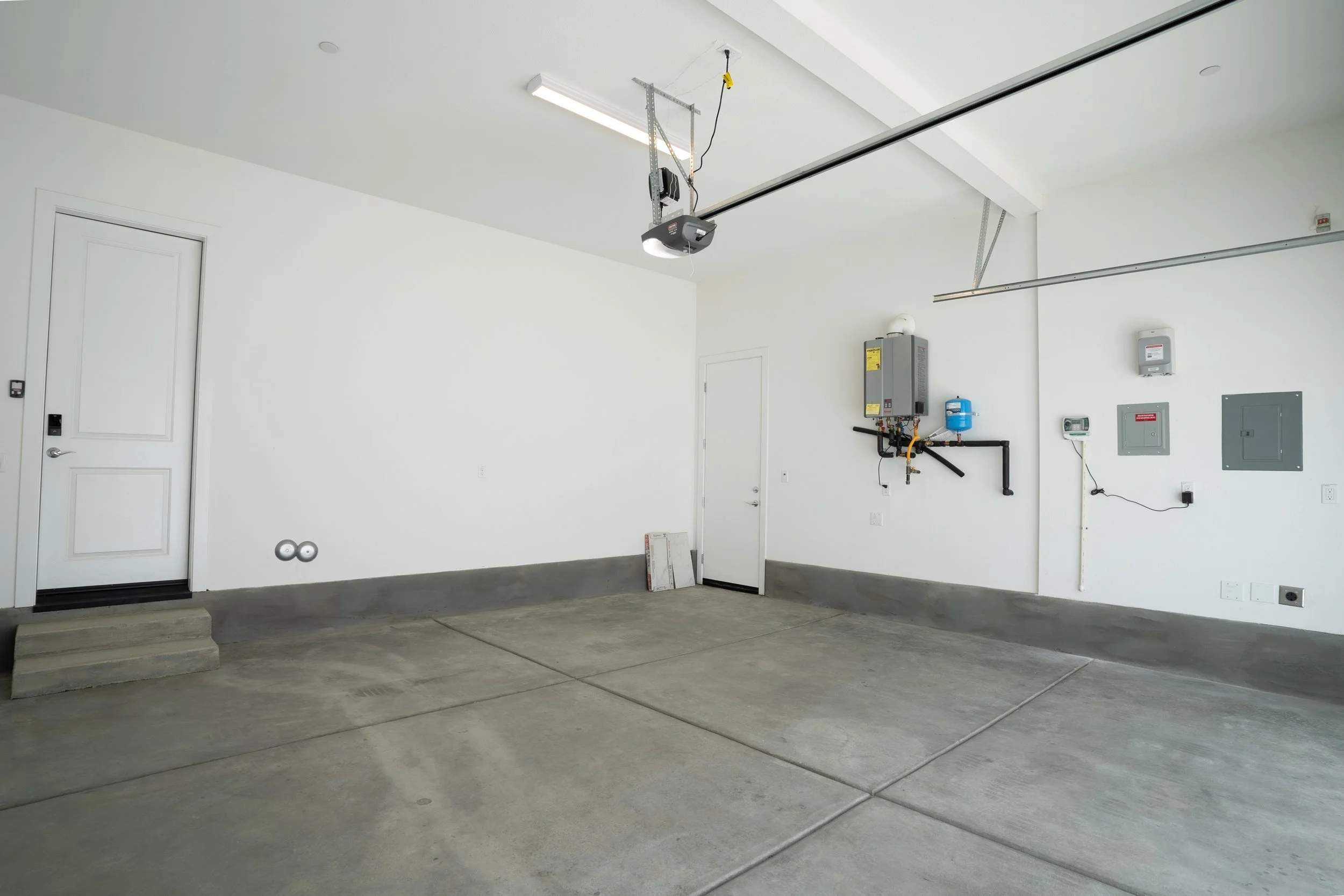 Empty garage with gray concrete floor, white walls, ceiling-mounted garage door opener, and utility panels on wall.