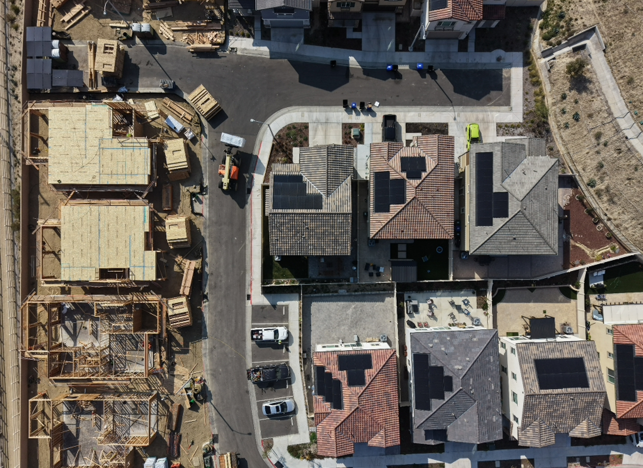 Aerial view of a residential neighborhood showing some houses under construction with wooden frameworks, neighboring completed homes with roofs featuring solar panels, a street with parked cars, and a small yard with tables and chairs.