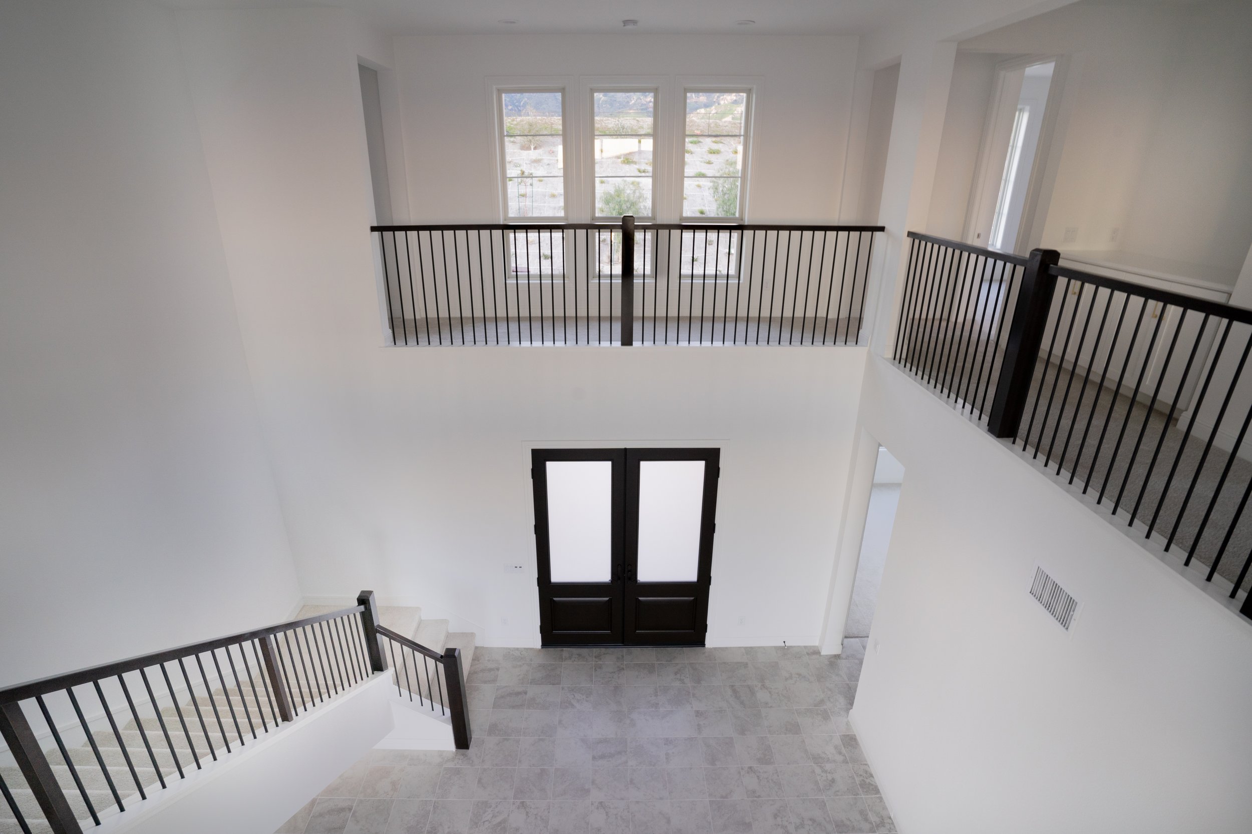 Interior view of a modern multi-story home entrance with a double door, tiled floor, white walls, and black metal railings on the upper level balcony overlooking the entryway.