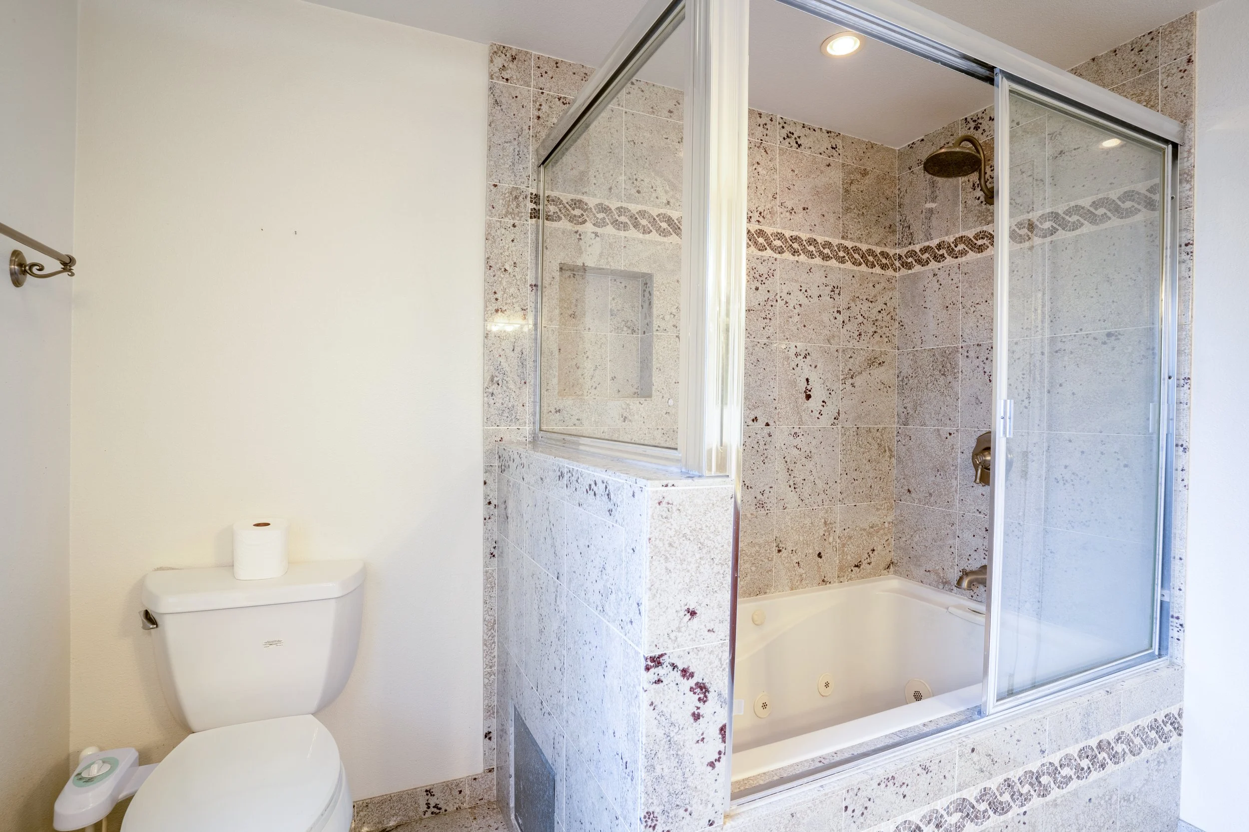 Bathroom with toilet and walk-in shower, cream-colored walls, and tiled shower with decorative band in brown and beige, cup toilet paper on toilet, and a mop next to it.