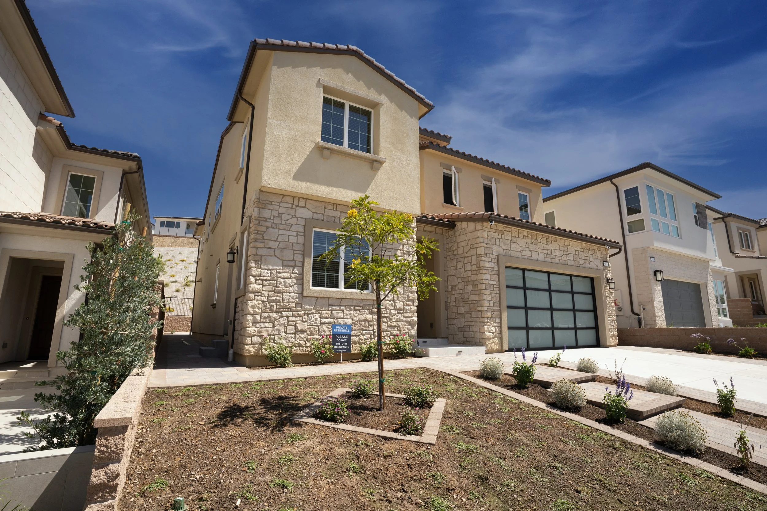 A modern multi-story house with beige and stone exterior, a garage with frosted glass door, small landscaped front yard with a young tree, potted plants, and a clear blue sky.