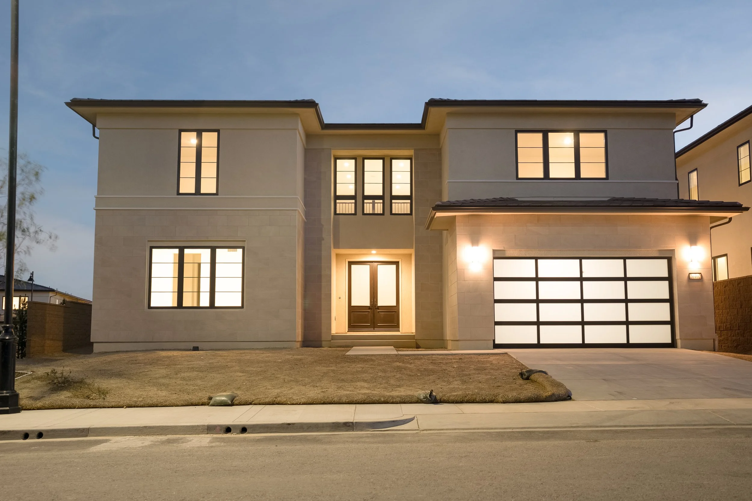 Modern two-story house with illuminated windows and garage door, front yard with grass and a sidewalk at dusk.