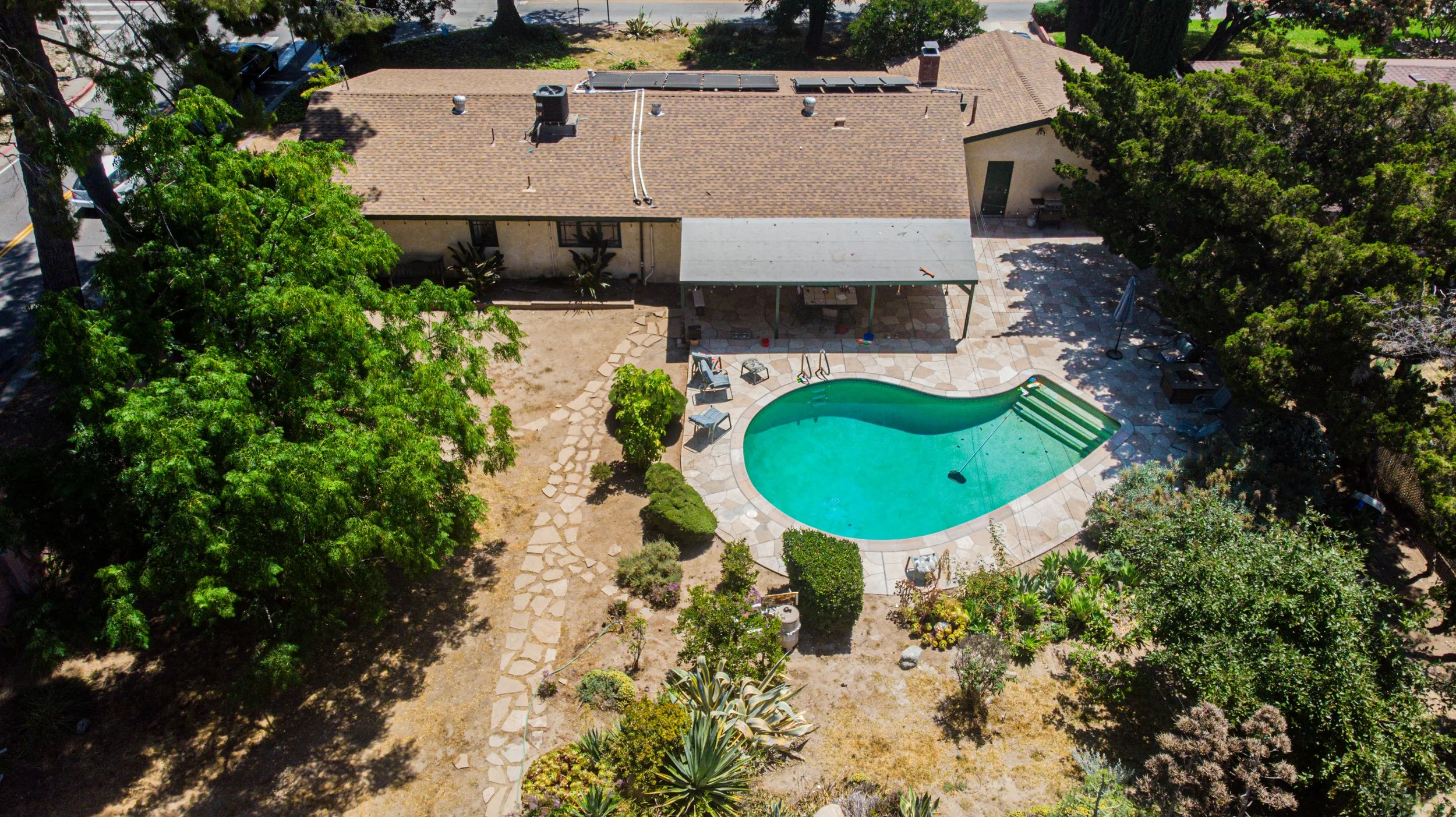 Aerial view of a backyard with a kidney-shaped swimming pool, surrounded by trees, plants, and a patio with chairs and umbrellas.