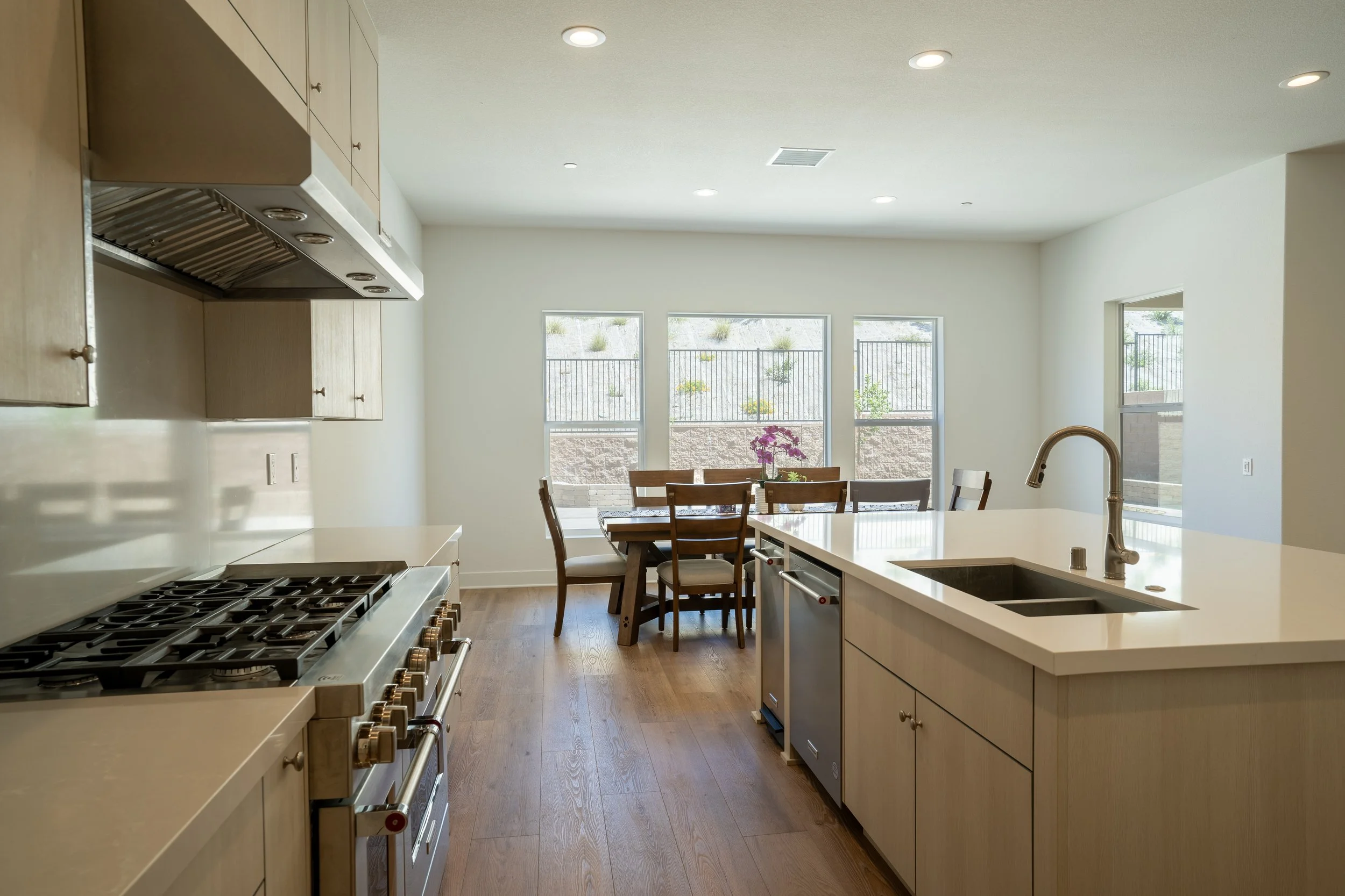 Modern kitchen with white countertops, stainless steel appliances, wooden cabinets, wooden dining table with six chairs, large windows with outdoor view, and hardwood flooring.