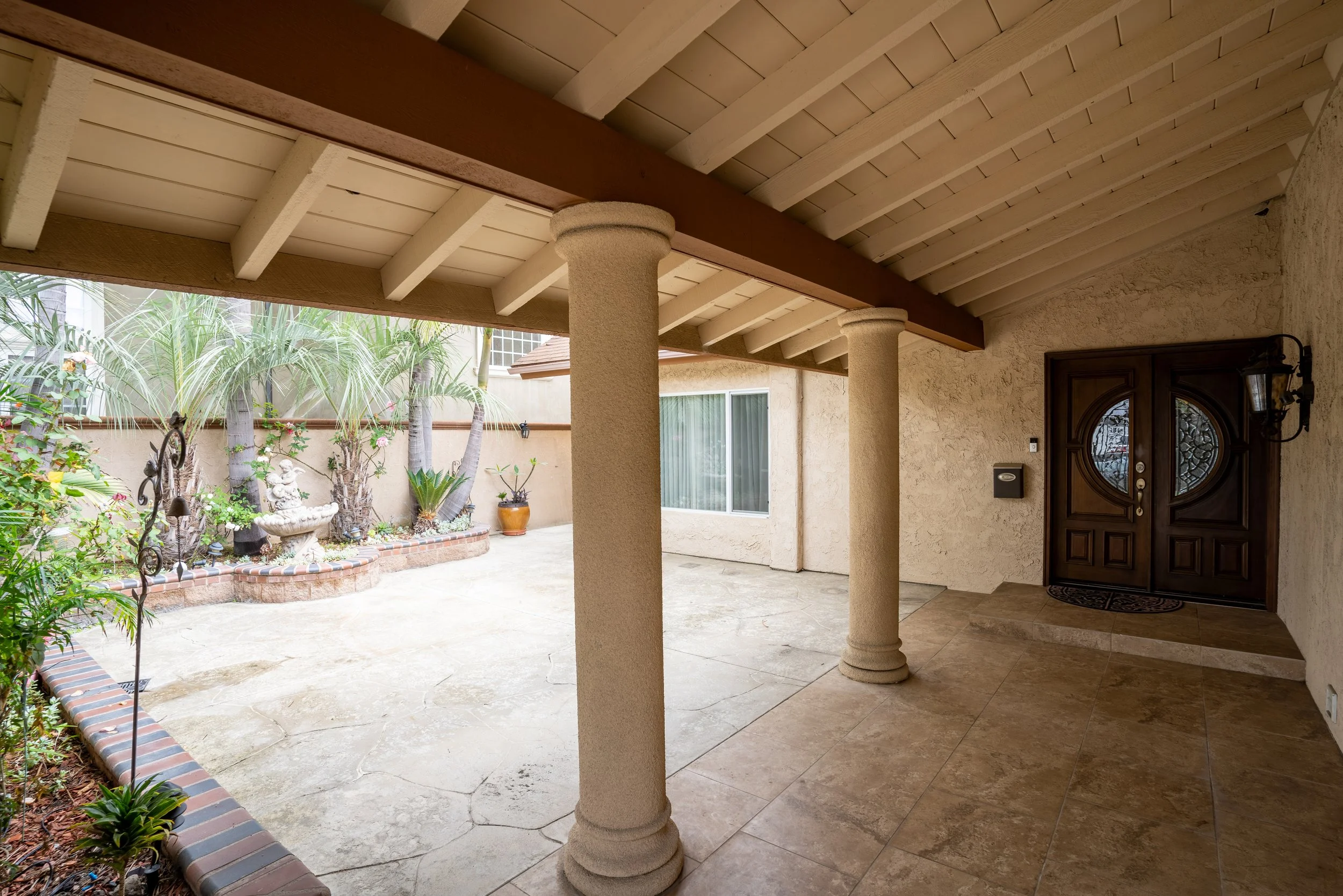 Covered front porch with beige stone columns, wooden ceiling, and tiled floor; view of a backyard with concrete patio, plants, trees, and garden statues; dark wooden front door with ornate glass panels.