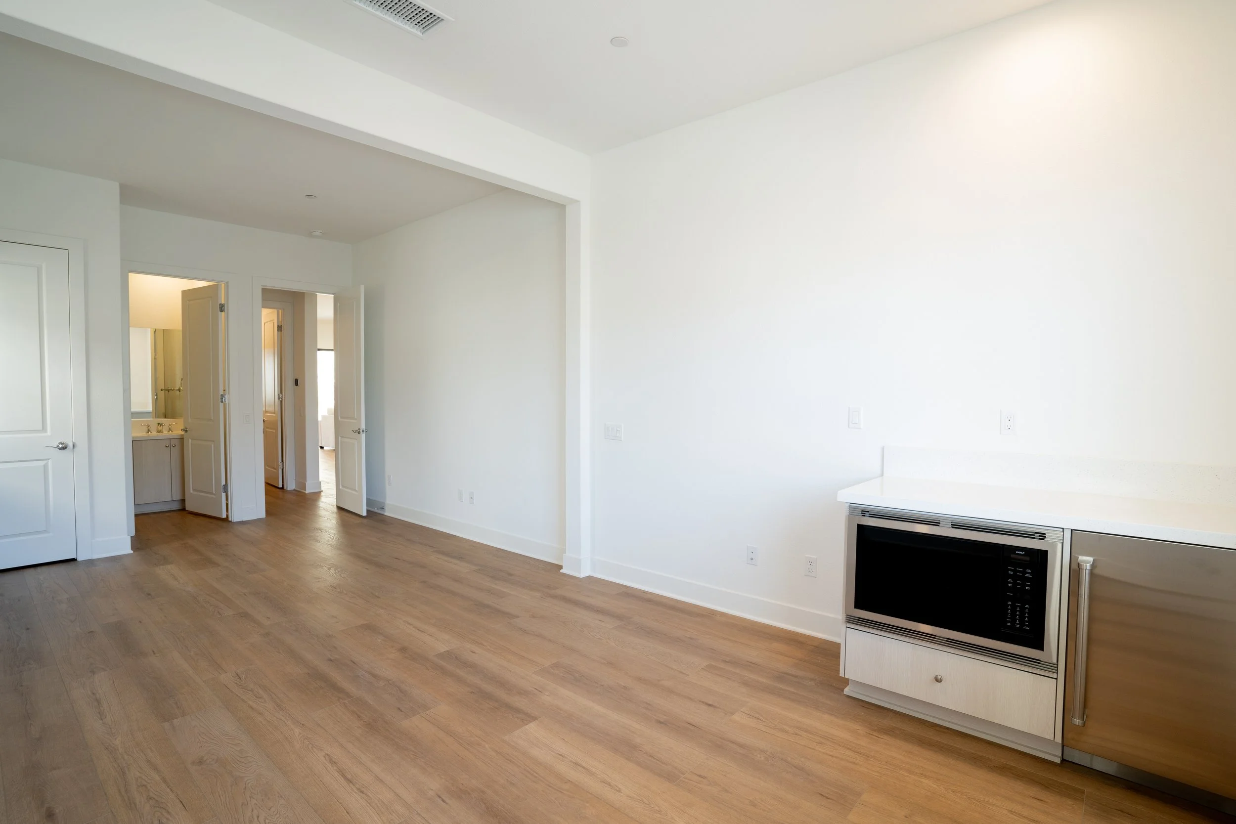 Empty room with white walls, wood flooring, and a built-in microwave oven, with doors leading to a bathroom and other rooms.