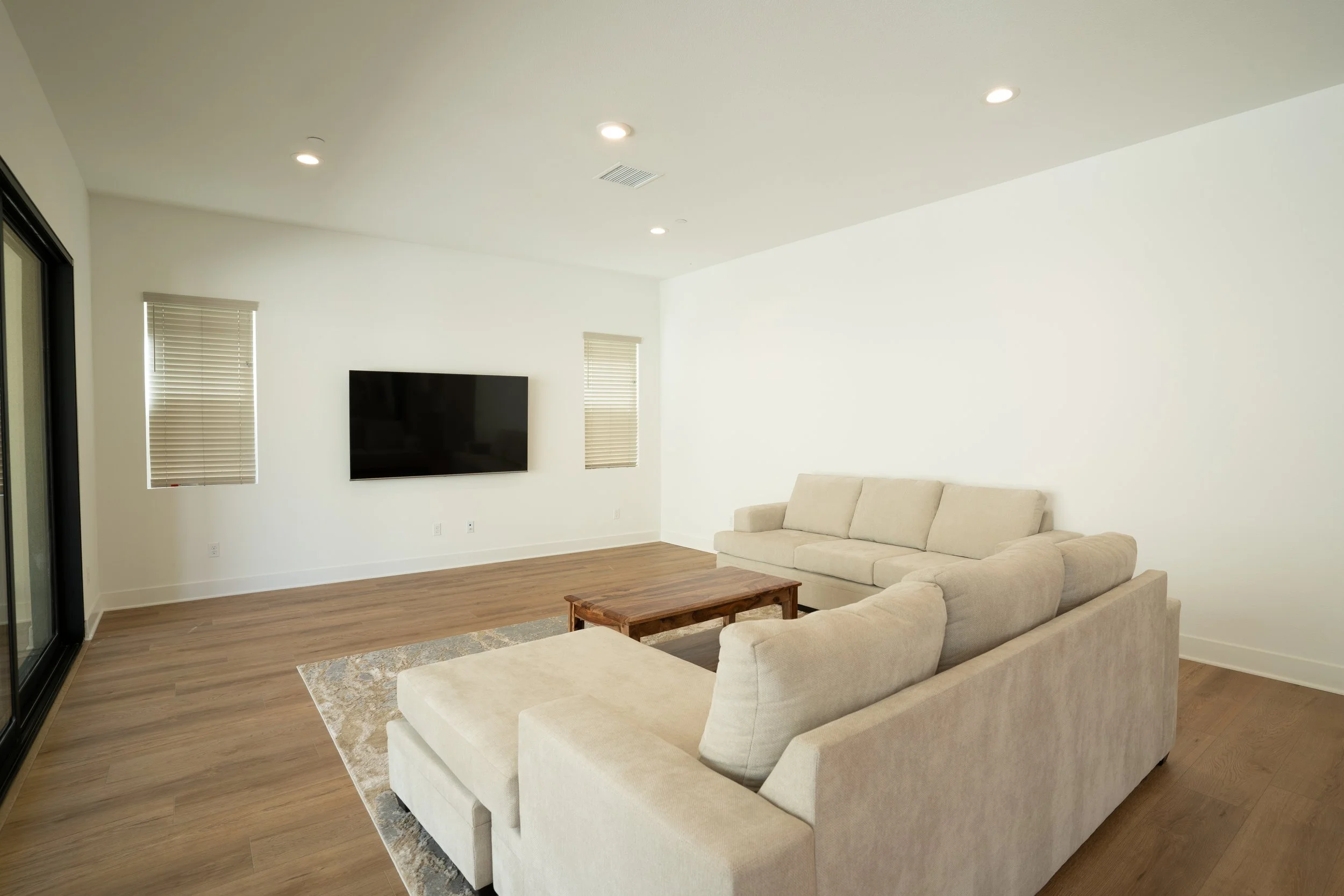 Empty living room with beige sofas, a wooden coffee table, a flat-screen TV on white wall, hardwood floors, and windows with blinds.