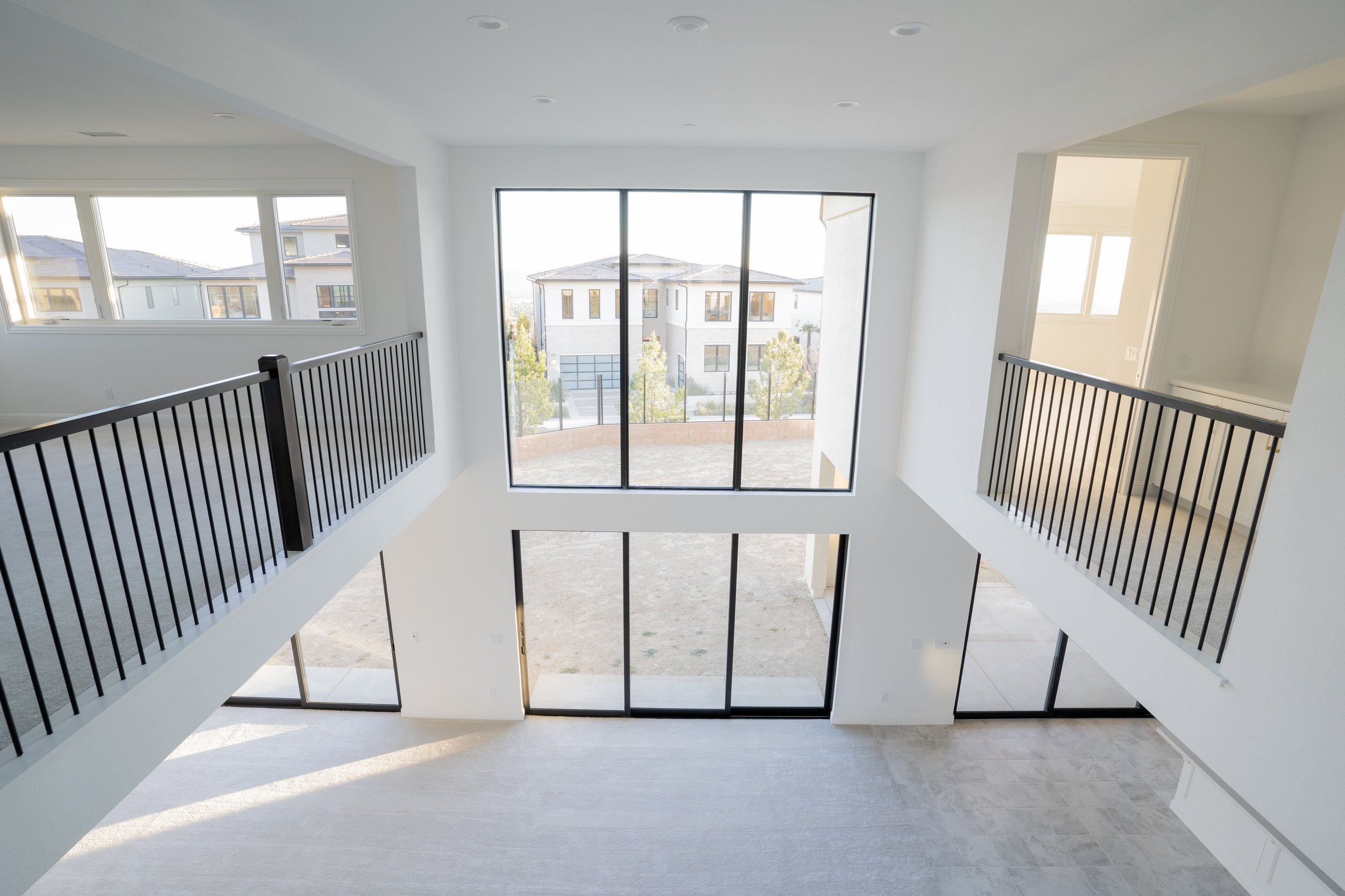 Empty, modern two-story house interior with large windows, white walls, black railings, and a view of neighboring houses outside.