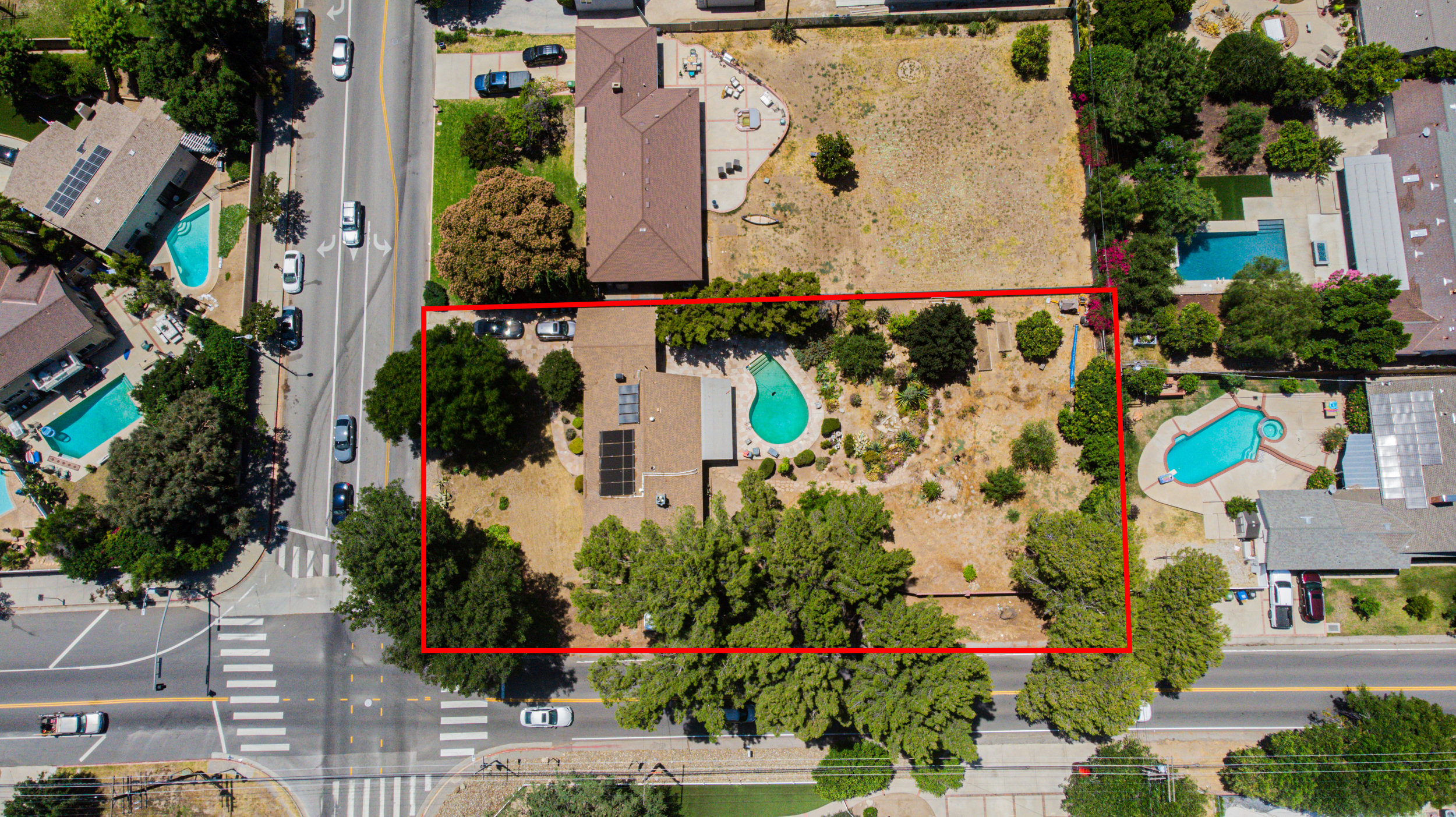 Aerial view of a residential neighborhood featuring multiple houses with swimming pools, surrounded by trees, with streets and parked cars, and a red outline marking a specific backyard with a small pool and garden.
