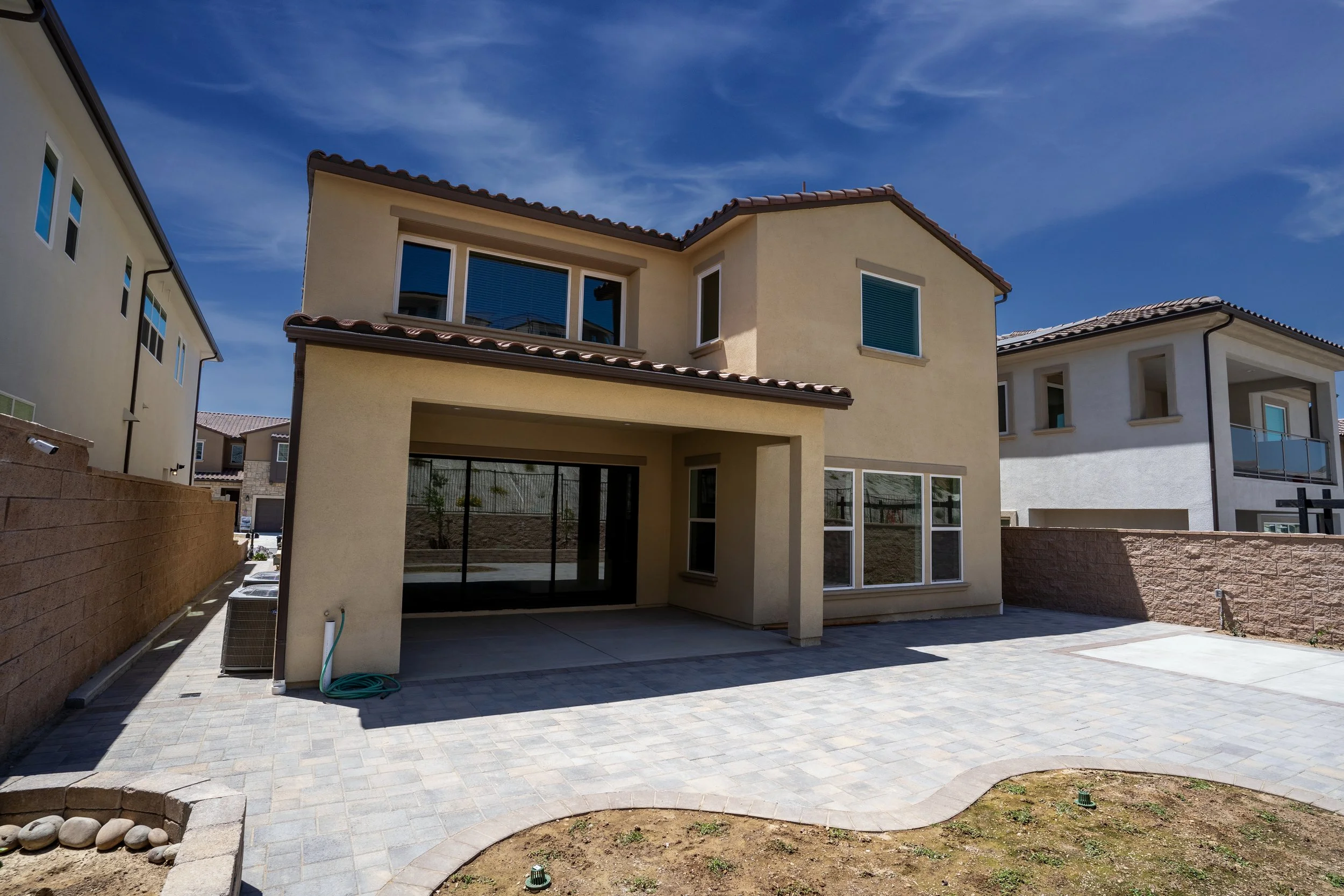 A two-story modern house with yellow stucco exterior and a tile roof, viewed from the backyard. The yard has a paved patio area and a small section of dirt in the foreground. The sky is bright blue with a few clouds.