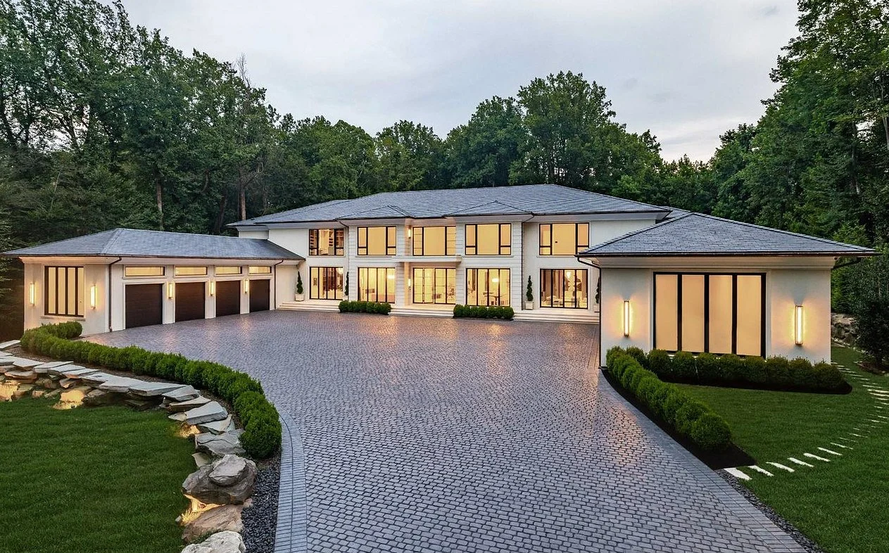 Large modern two-story house with white exterior, multiple large windows, attached garage, and a spacious paved driveway surrounded by landscaped greenery.