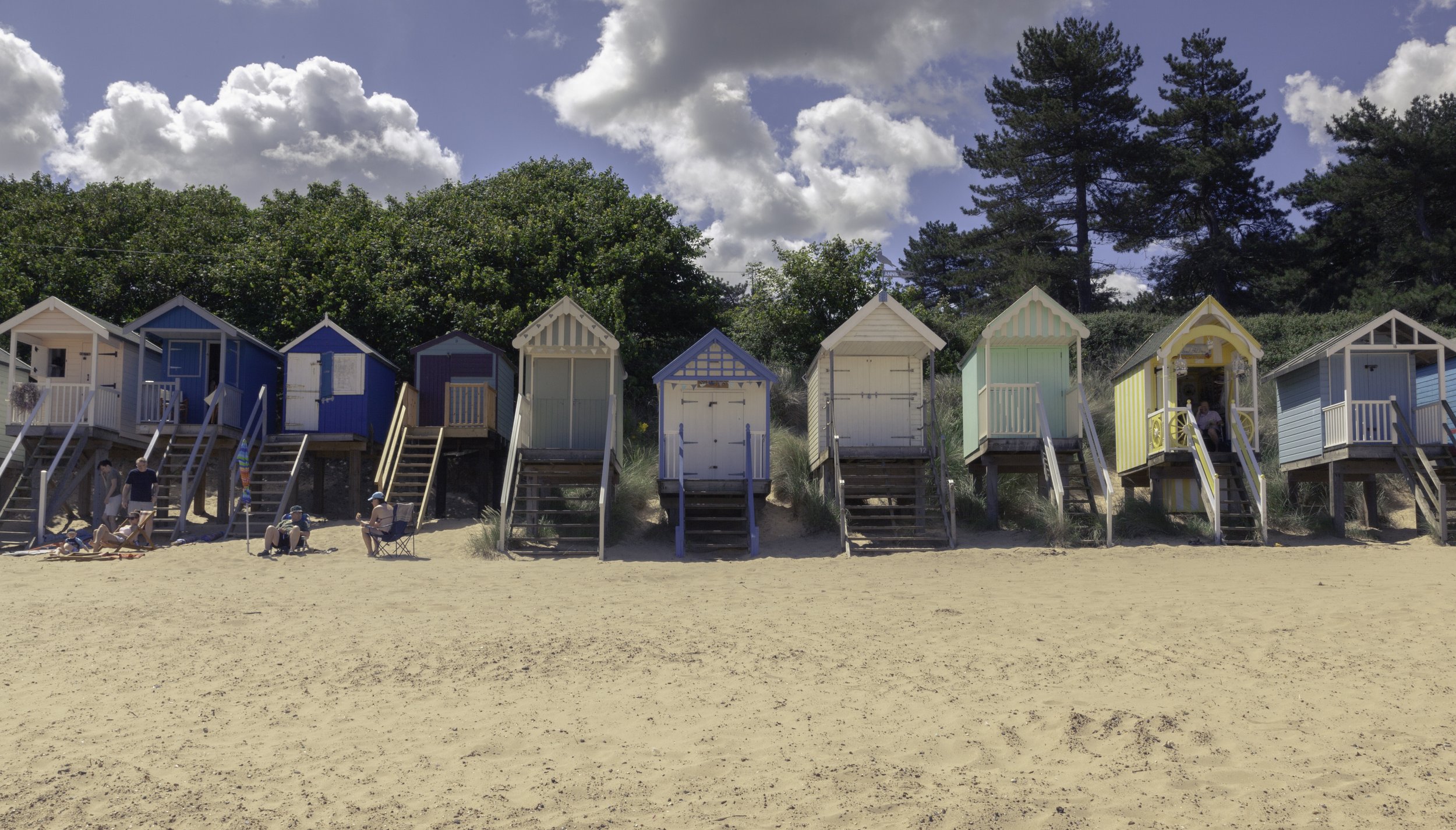 Colorful beach huts on stilts lined up along sandy shore, with people sitting and relaxing, green trees and a partly cloudy sky in the background.