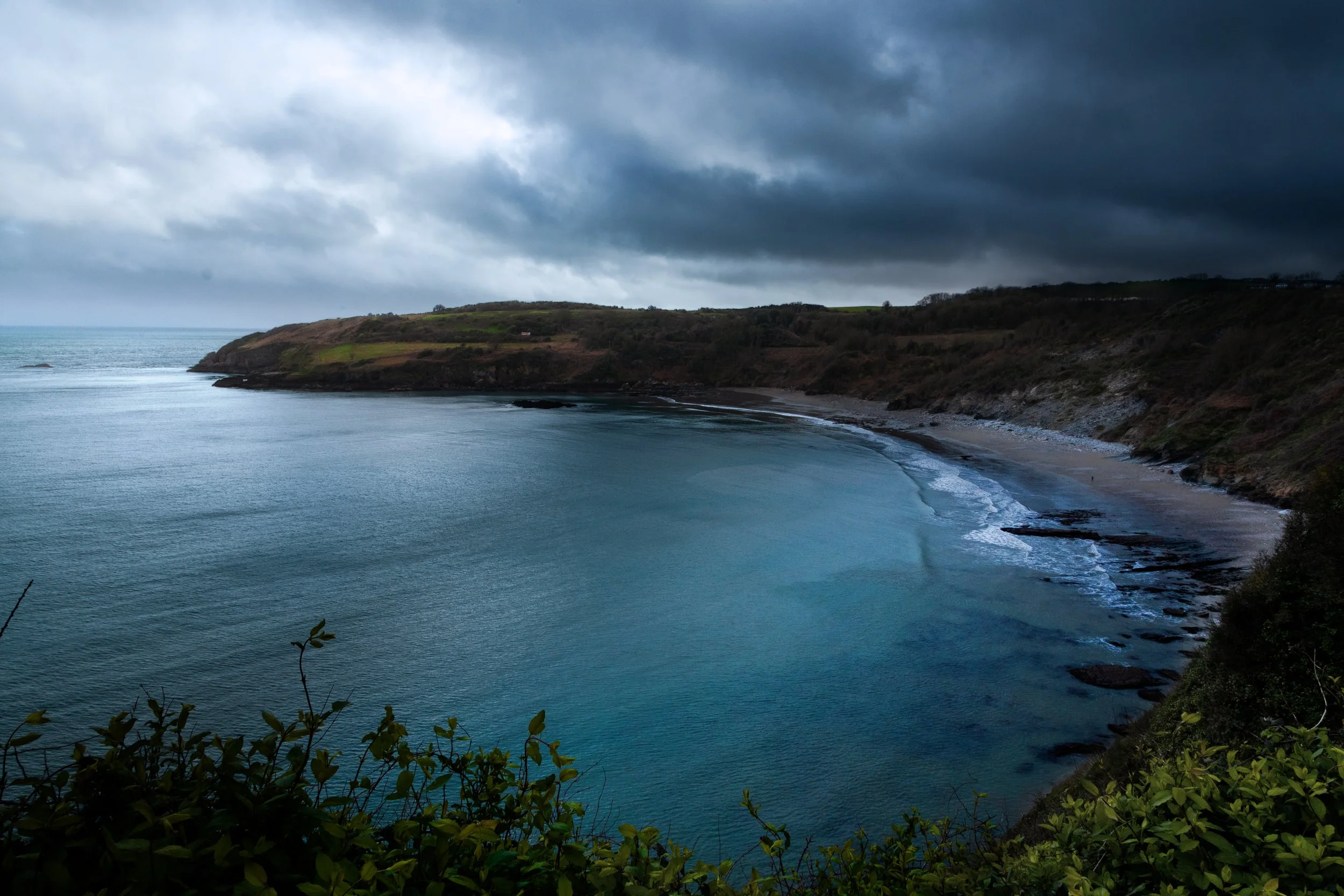 A coastal landscape on a cloudy day, with a curving sandy beach, rocky shoreline, green-hilled cliffs, and dark storm clouds overhead.