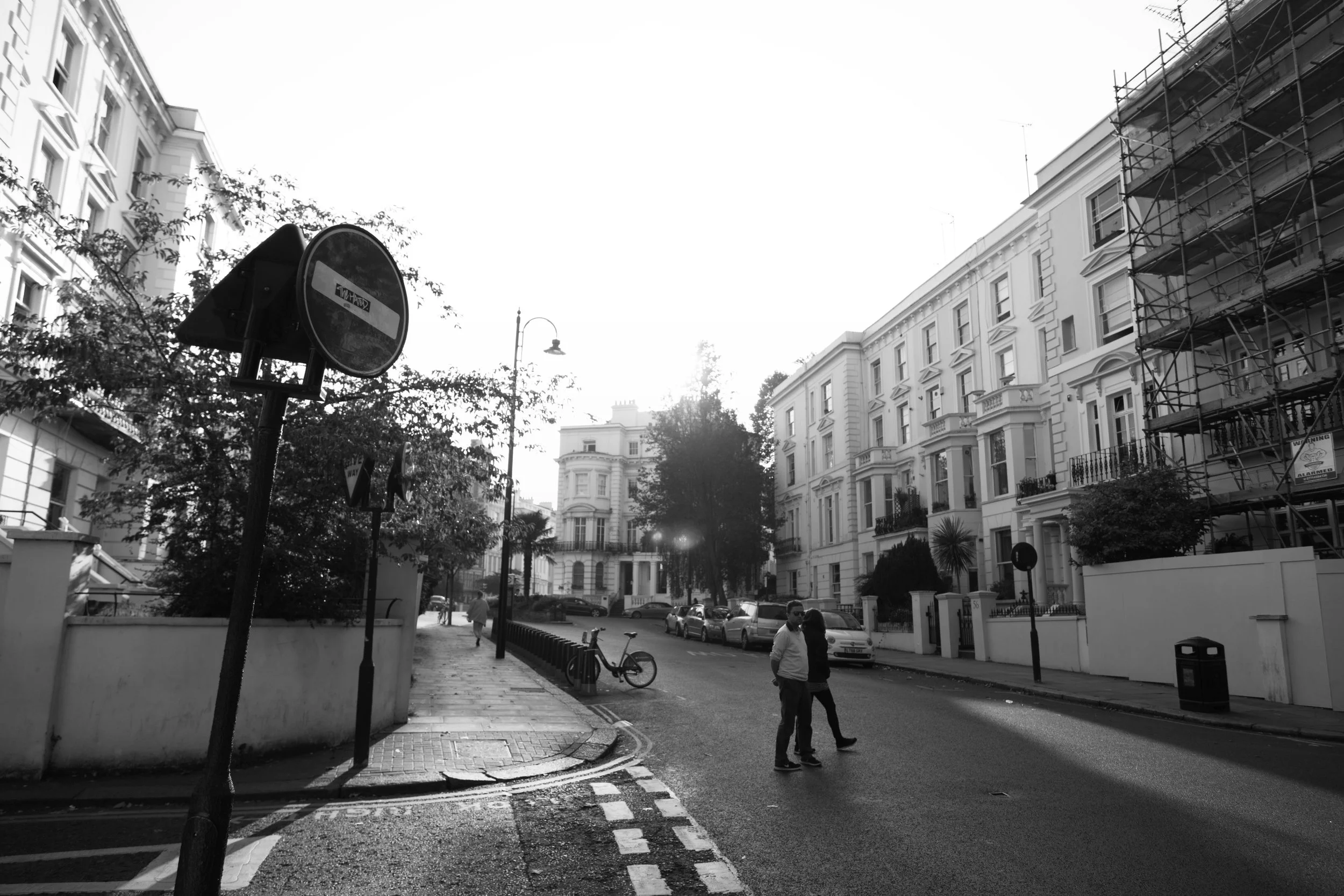 Black and white photo of a city street with buildings, parked cars, and people crossing the road, with a bicycle leaning against a wall and a 'no entry' sign on a pole.