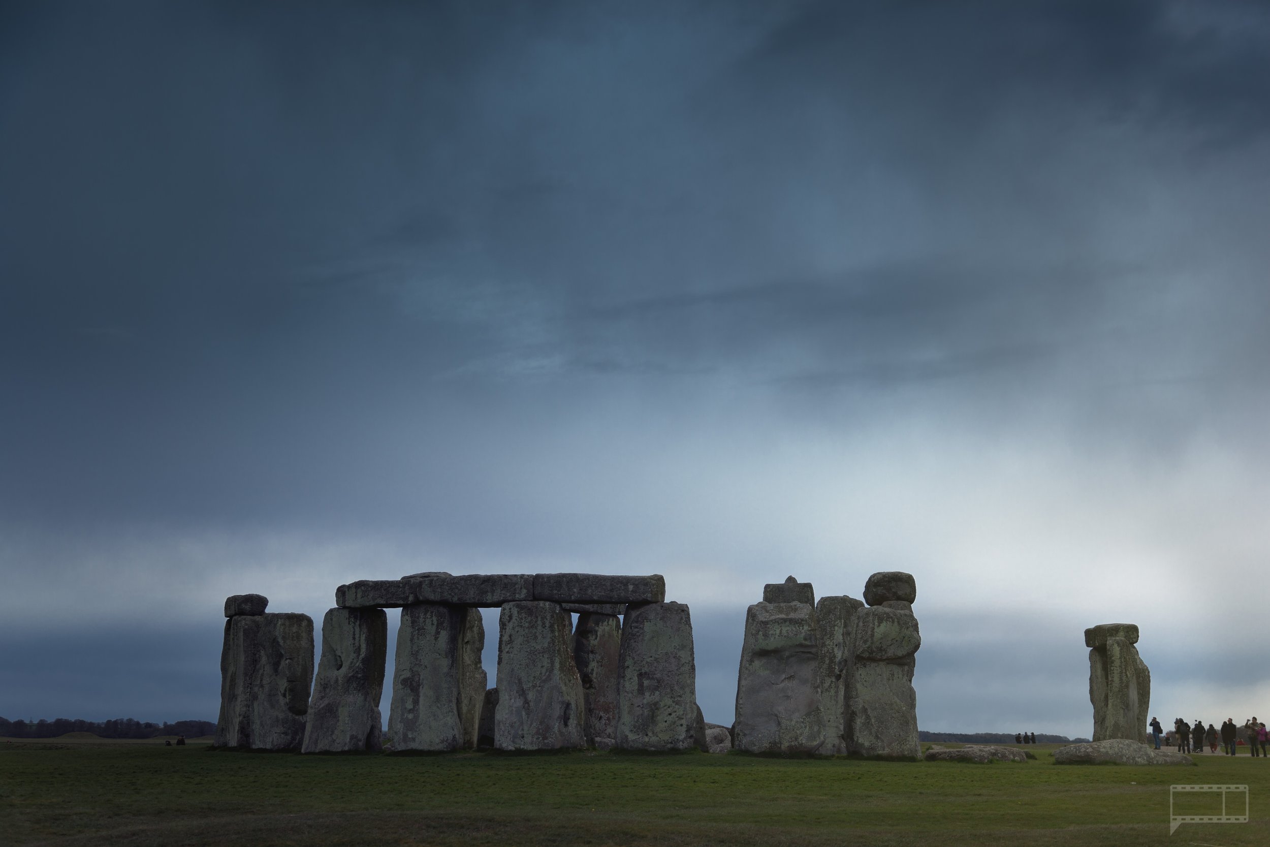 Stonehenge with a cloudy, dark sky and a group of people on the right side.
